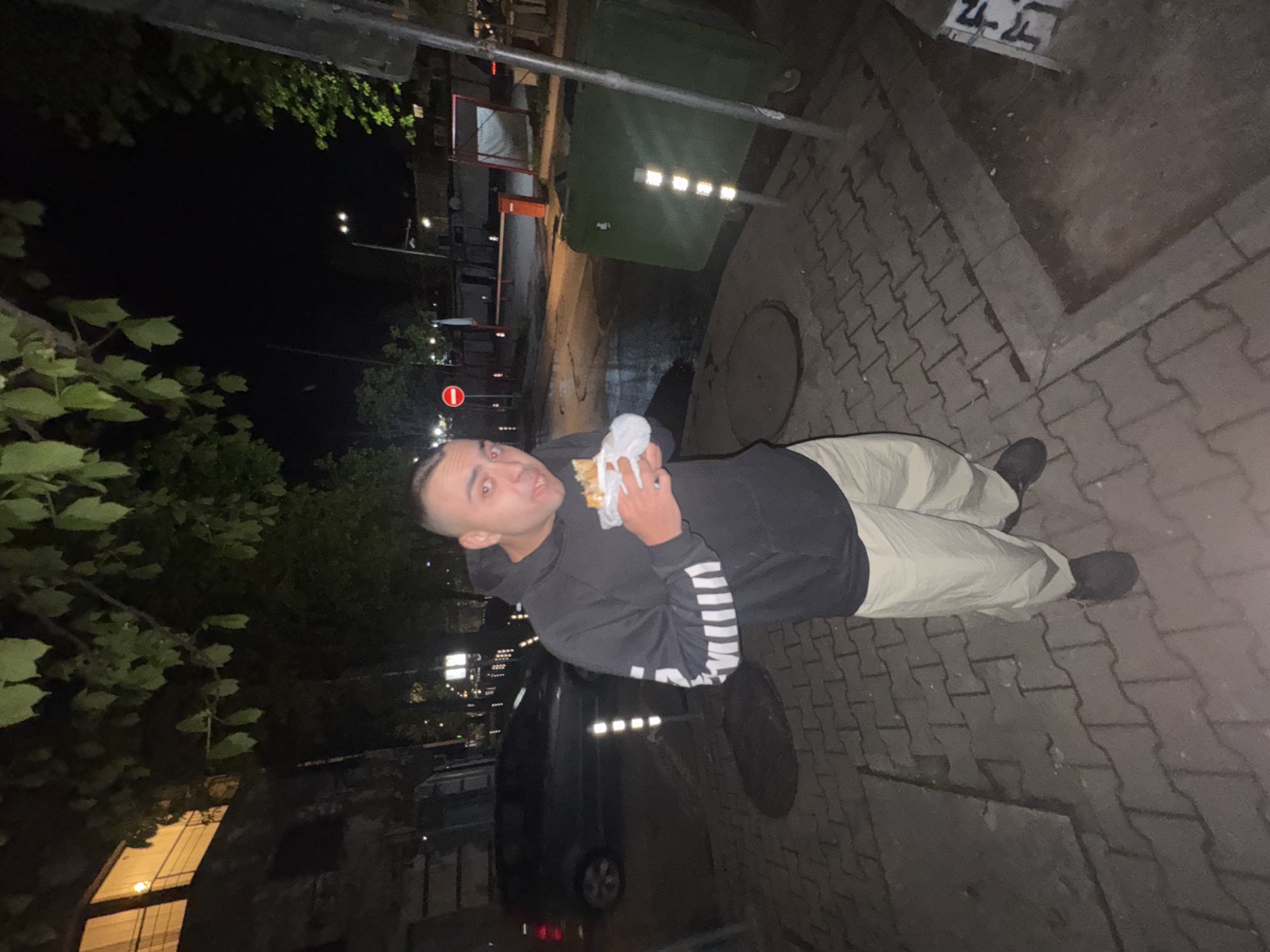 A man stands on a brick sidewalk in Sofia, Bulgaria. He is holding a wrapped sandwich and looking at the camera. He is wearing a black hooded sweatshirt with white stripes on the sleeves and khaki pants, and dark shoes. A car is parked behind him. A garbage bin, a manhole cover, a street, a traffic sign and trees are visible in the background. Some buildings are visible in the distance. It is night.