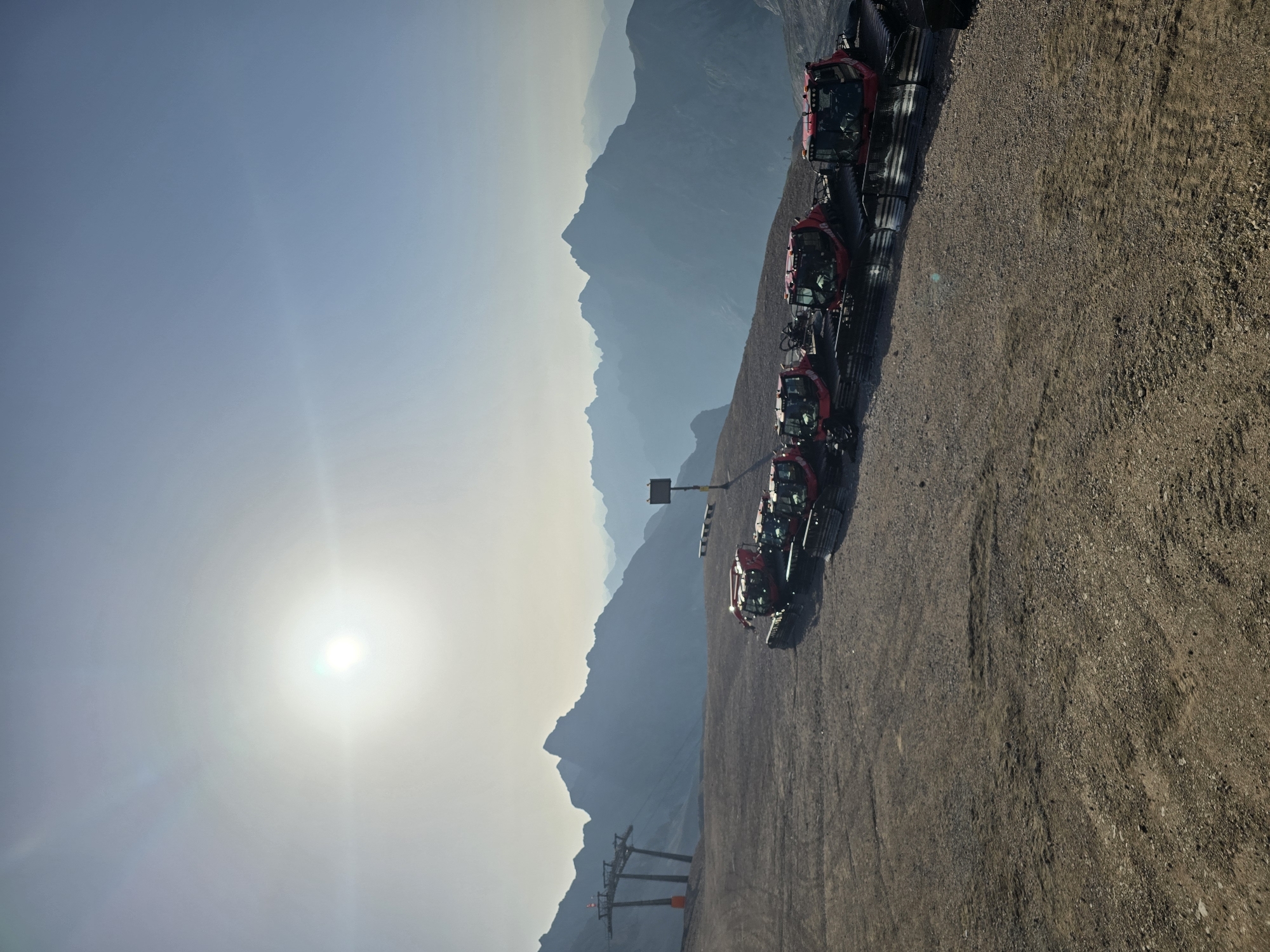 The image captures a high-altitude mountain scene, likely at the Zugspitze, the highest peak in Germany, near Garmisch-Partenkirchen. The foreground features a dusty, gravelly slope with several bright red snow groomers lined up. These machines are equipped with tracks and plows, indicating preparation for winter conditions. In the mid-ground, a ski lift structure with towers and cables is visible on the left, suggesting a ski resort setting. A yellow signpost stands near the center of the frame. The background is dominated by a vast, hazy mountain range with layers of peaks receding into the distance. The sky is a clear, pale blue with a bright, slightly diffused sun low in the sky, casting a soft light over the scene. There are no people visible in the image.