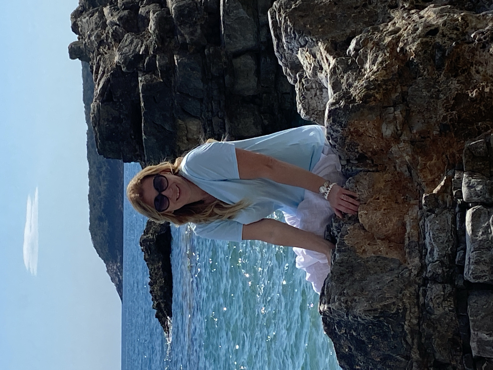 A medium shot, rotated 90 degrees counter-clockwise, shows a woman in a light blue top and white skirt nestled between rocks on a sunny day. The location is likely Prijevor, Montenegro, as the image appears to be from that area. The woman has blonde hair and is wearing sunglasses. She is smiling and appears to be posing for the photo. In the background is blue water and a rocky coastline with green trees. The sunlight reflects off the water creating bright, shimmering highlights.