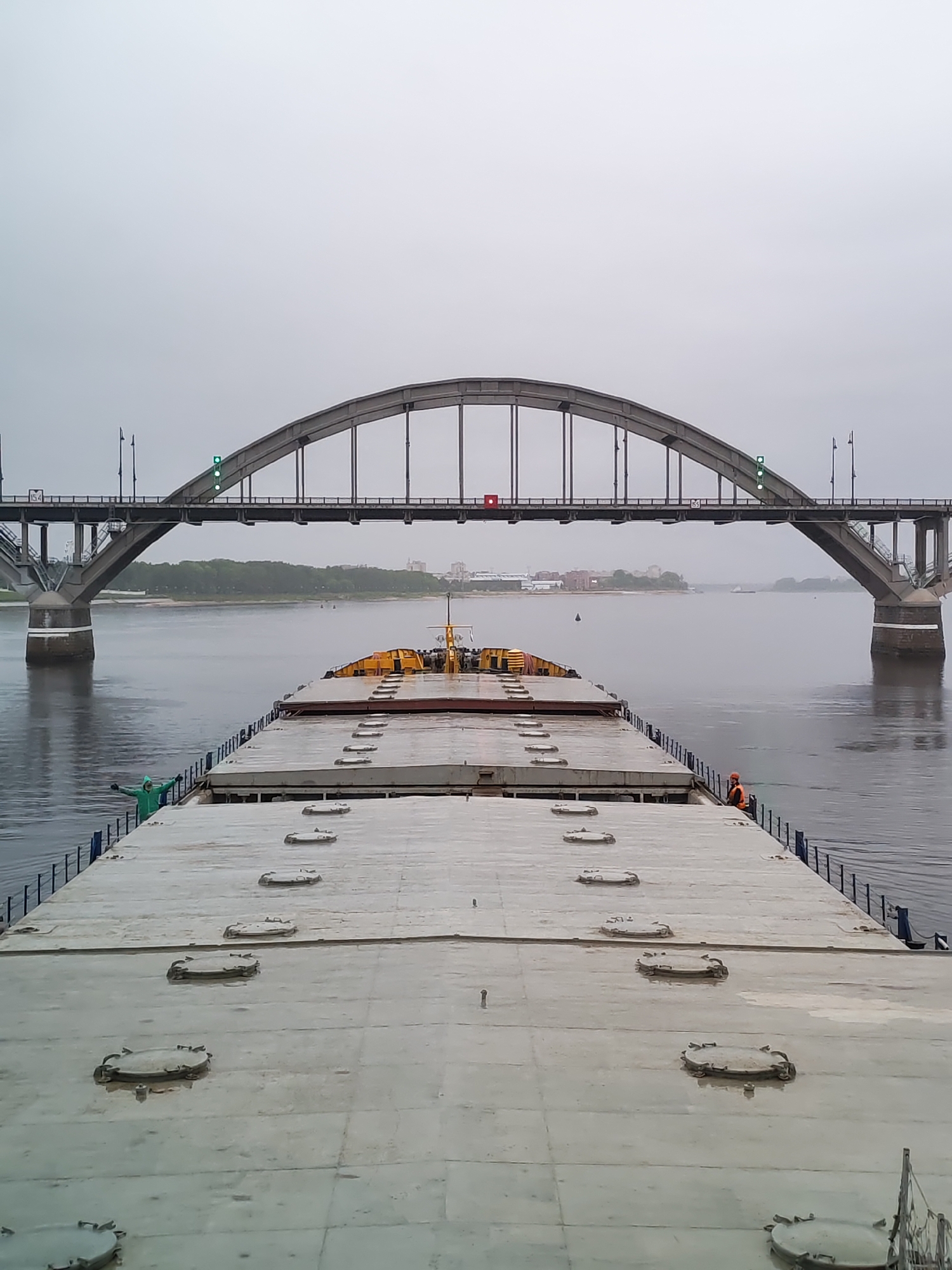 The image depicts a view from the deck of a barge, potentially on the Volga River near Bol'shie Mkhi, Russia. A large arched bridge spans the river in the background. The barge has a flat, gray deck with multiple circular hatch covers visible. Two individuals are visible on the barge's edges, one in green and the other wearing orange. The environment is overcast, with a dull, gray sky. The river's surface is calm, and some greenery is visible along the banks in the distance.