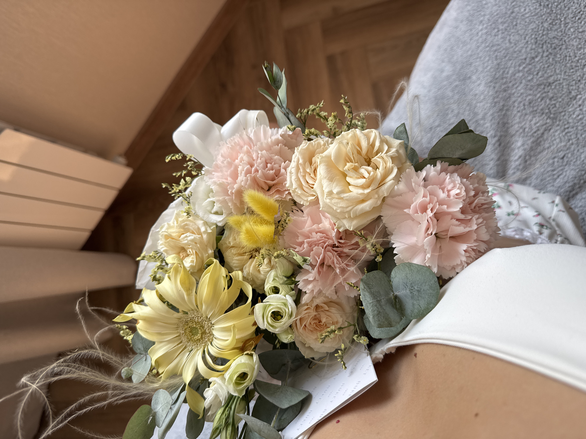 The image shows a bouquet of flowers held against a backdrop of a person's upper chest and shoulder area. The bouquet features a mix of pastel colors: soft pink carnations, creamy yellow roses, and a large yellow daisy-like flower. Green eucalyptus leaves and other decorative foliage are interspersed among the blooms. A white ribbon is visible, adding a touch of elegance to the arrangement. The setting appears to be indoors, with glimpses of wooden flooring and neutral-colored upholstery in the background. The location is Unknown, Unknown.