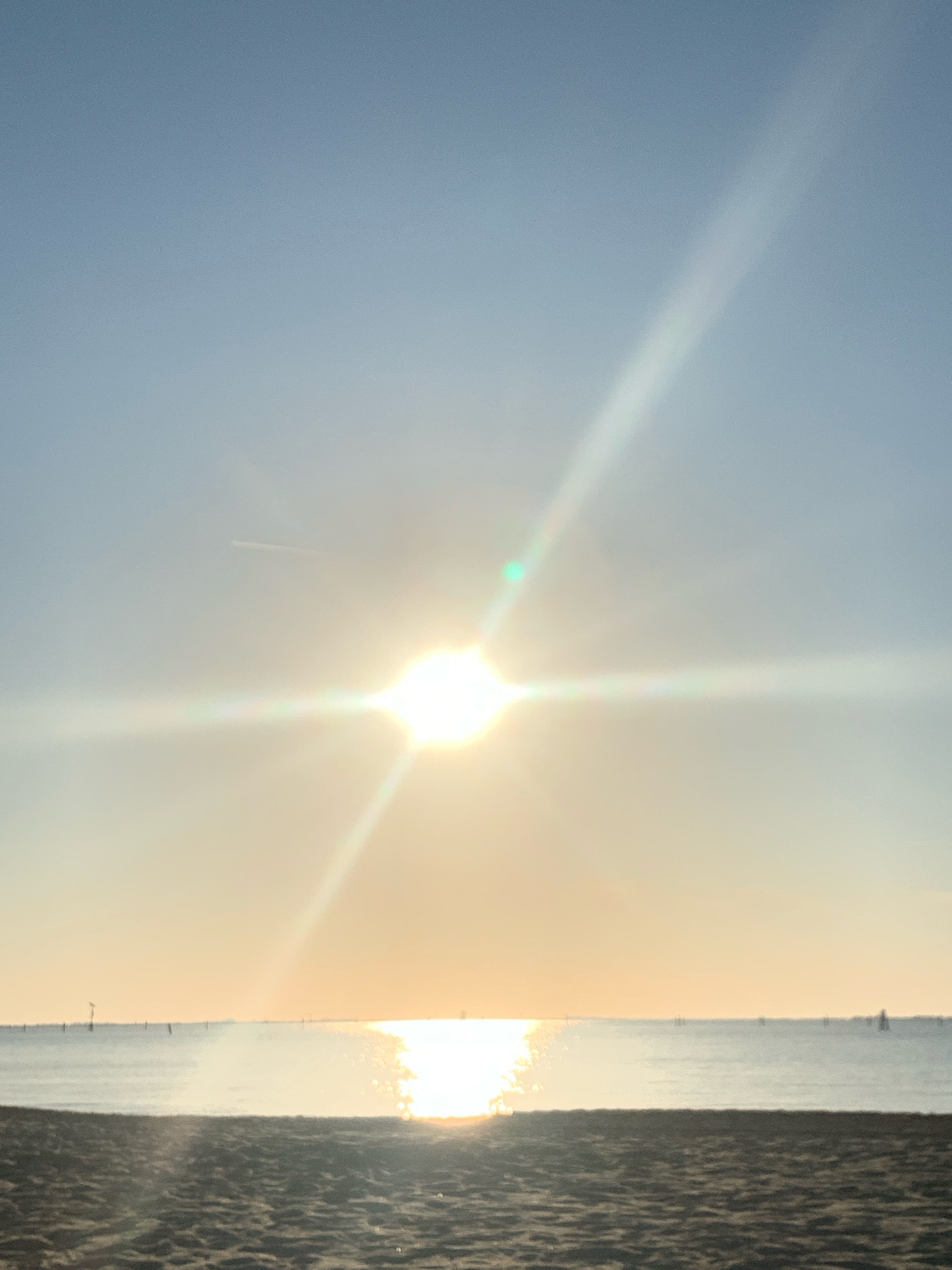 The image depicts a serene beach scene at dawn or dusk, likely overlooking the Adriatic Sea. In the foreground, the coarse texture of the sand is visible. The middle ground is dominated by the calm, shimmering water, reflecting the bright sun. Distant, indistinct objects, possibly buoys or small boats, are scattered across the horizon line. The sky transitions from a pale blue at the top to a warm, golden hue near the horizon, where the sun is a brilliant, starburst-like presence, casting long rays and lens flare across the scene. A faint contrail from an aircraft is visible in the upper portion of the sky. The overall impression is one of tranquility and natural beauty. Given the reference to Comacchio, Italy, this scene could be situated along its coastline, which features extensive beaches and lagoons.