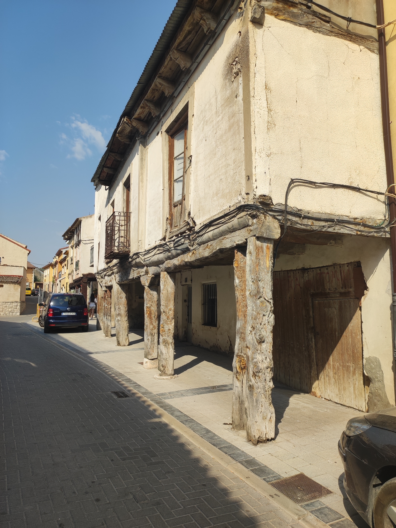 This is a street-level, eye-level, medium shot of a rural Spanish town, likely Traspinedo, Spain, on a sunny day. The focus is on a weathered building with a covered walkway supported by thick, rough-hewn wooden posts. The building's facade is rough plaster, light beige in color, with visible cracks and signs of age. The roof has a corrugated metal covering.
The walkway is paved with light-colored tiles, bordered by a darker tiled curb. Electrical wires are strung along the building's exterior, running above the wooden support beams and under the roof. A dark wooden double door is visible on the right side of the building, set within the covered area. To the left of the wooden posts, a small, barred window is present.
In the street, a dark blue car is parked, facing away from the camera. The street itself is paved with dark gray interlocking bricks laid in a herringbone pattern. The sky is clear blue with a few wispy white clouds. The overall impression is one of an old, rustic building in a quiet, sunny Spanish town.