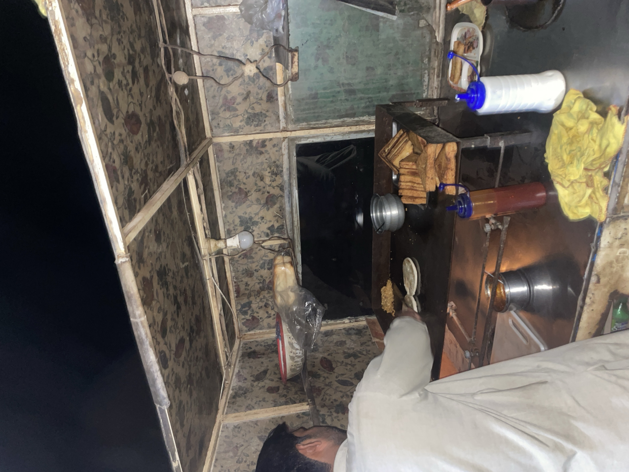 This image depicts a nighttime scene at a small, rustic food stall in Karamsar, Pakistan, where a person is actively preparing food. The stall is characterized by its metal frame and floral-patterned panels, giving it a makeshift or traditional appearance.
The primary subject is a person, visible from the torso up, wearing a white tunic (likely a shalwar kameez top) and a beard. They are holding a spatula or ladle over a flat cooking surface, possibly a griddle or tawa, where ingredients are being cooked or heated.
Various objects related to food preparation and service are arranged on the counter. These include stacks of sliced bread, a white plastic container holding what appear to be fried snacks (possibly spring rolls or similar items), and two squeeze bottles with blue caps—one white (likely for mayonnaise or cream) and one amber-colored (likely for a sauce like ketchup or chili). A yellow cloth or rag is crumpled on the counter.
The scene is illuminated by a bare light bulb hanging from a wire, suggesting limited artificial lighting. The background outside the stall is completely dark, confirming that the activity is taking place at night. A small green bottle, possibly containing a beverage, is partially visible on the right side of the counter. The overall setting is indicative of a local street food vendor or a small, informal eatery. No discernible text is clearly visible on any objects.