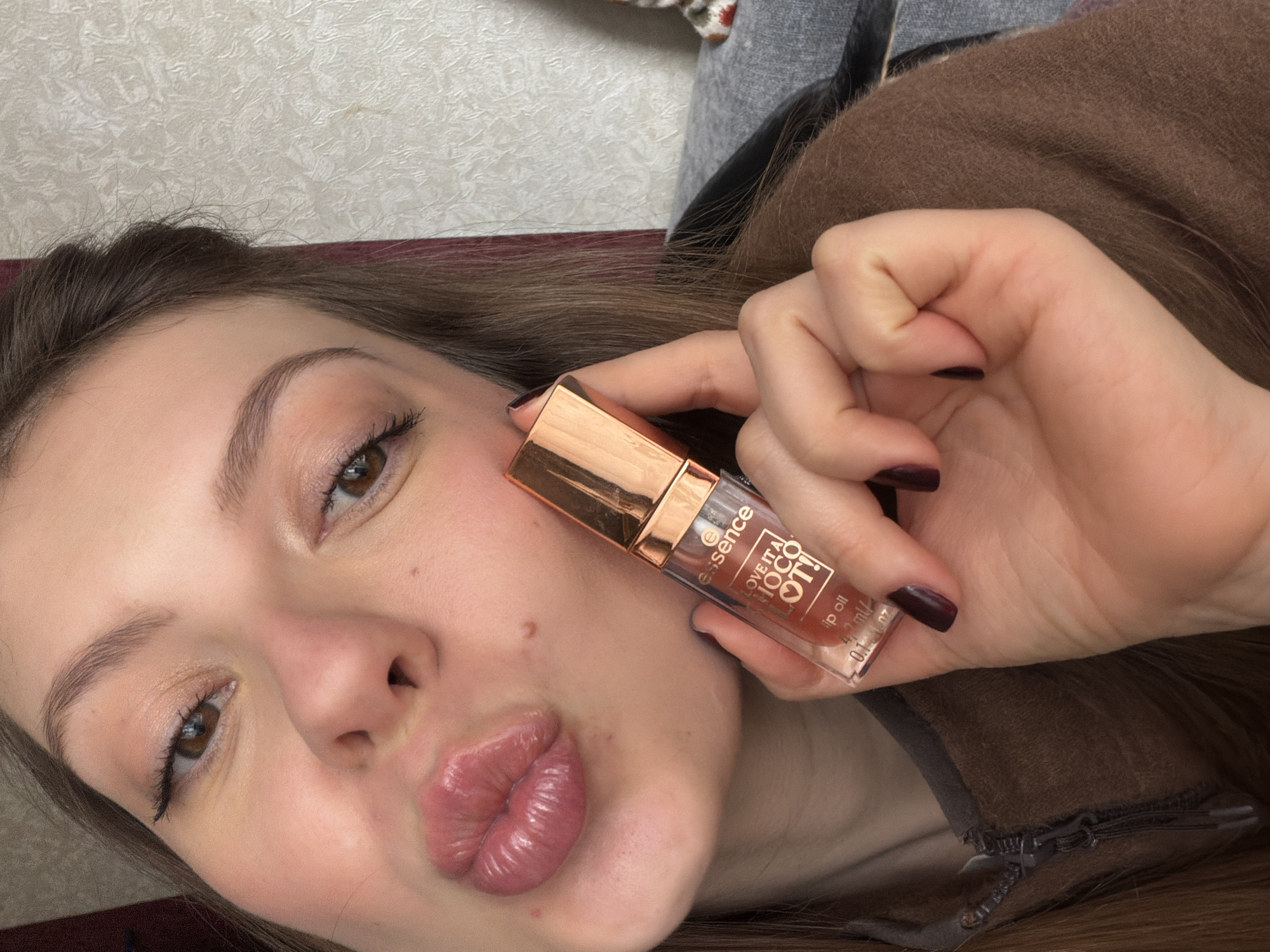 The image features a close-up, diagonal shot of a young woman with brown hair and fair skin. She is looking towards the camera with a playful expression, her lips puckered in a glossy "kiss face." Her eyes are brown, and she has visible eyeliner and mascara, along with well-groomed eyebrows.
In her right hand, she holds up a small bottle of "essence LOVE IT! COCO! lip oil." The clear bottle has a rose gold cap and contains a brownish-pink liquid. Visible text on the label includes the brand "essence," the product name "LOVE IT! COCO!," "lip oil," and the volume "4.2 ml / 0.14 fl. oz." Her fingernails are painted a dark, possibly burgundy, color.
The woman is wearing a brown garment, possibly a sweater or jacket, over a grey layer. The background consists of a light-colored wall with a subtle, textured pattern. The scene is lit by what appears to be bright indoor lighting. The location context provided for this image is Khimki, Russia.