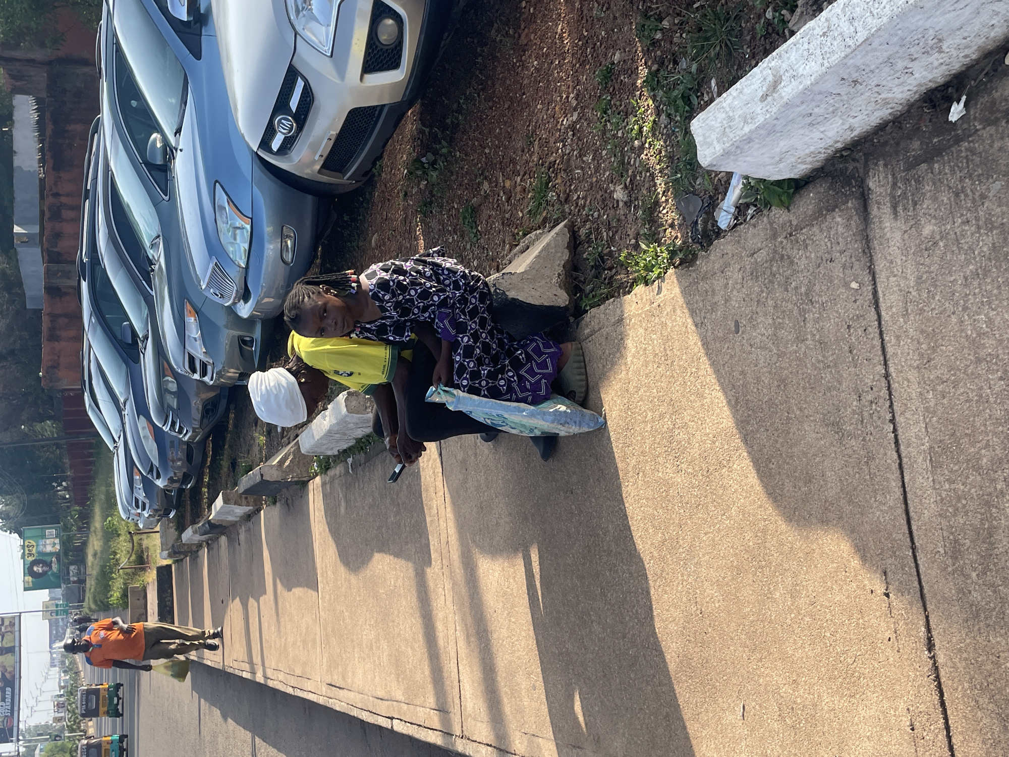 A man in an orange shirt and khaki pants walks along a concrete sidewalk next to a street lined with parked cars. He is carrying a plastic bag in his left hand. Two people, a woman wearing a white headscarf and a yellow shirt, and a young girl in a black and purple dress, are sitting on a concrete curb near the parked cars. The woman is looking at her phone. A blue and white patterned bag is between them. The scene appears to be in Jos, Nigeria, suggested by the style of the vehicles and the general environment. The sun is casting long shadows, indicating it is likely late afternoon or early morning.