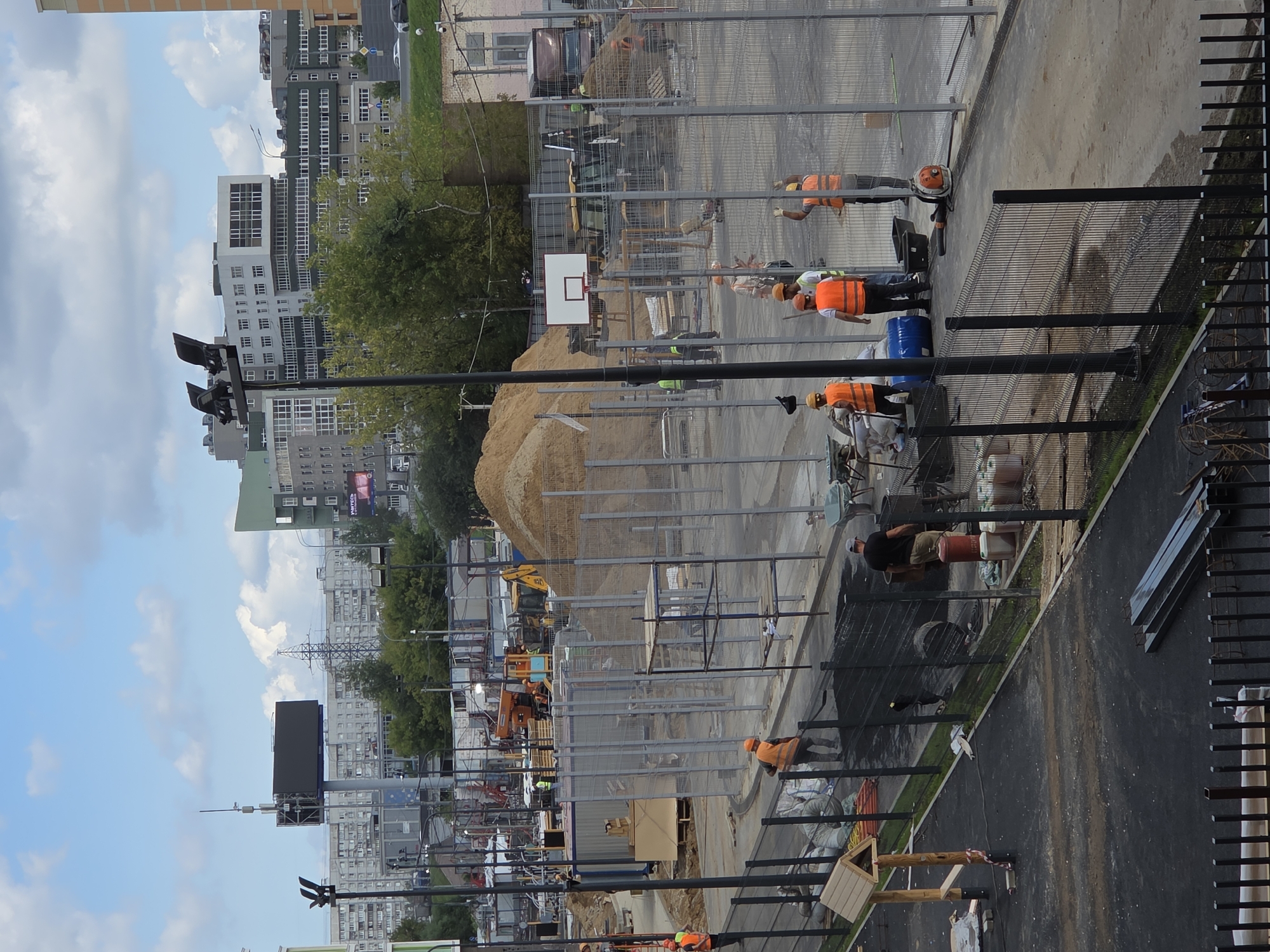 This image depicts a construction site in Moscow, Russia. A group of construction workers in orange safety vests and hard hats are visible, engaged in various tasks. In the foreground, a worker with a shovel is working on the ground. Further back, other workers are gathered near a blue barrel, and one appears to be operating a leaf blower. Construction materials, including sand, bags, and scaffolding, are present throughout the scene. In the background, a basketball hoop stands within the fenced-off area. The surrounding environment includes modern buildings, trees, and a cloudy sky. A large digital billboard is visible in the upper left quadrant of the image.