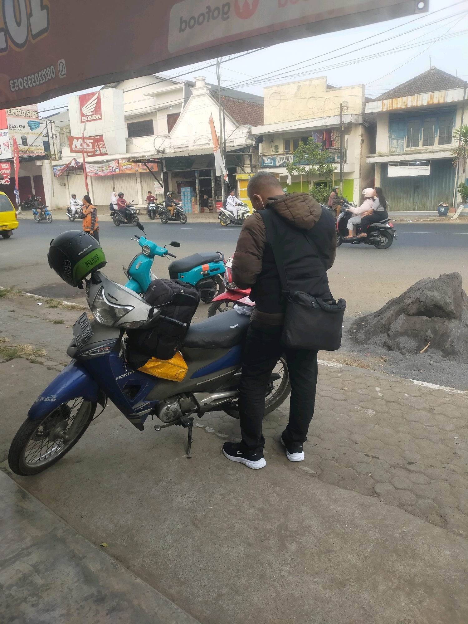 The image depicts a street scene in Bululawang, Indonesia, with a focus on a man standing next to a blue Honda motorcycle. The man, seen from behind, is wearing a brown hooded jacket, black pants, and black and white sneakers. He has a black messenger bag slung across his chest. The motorcycle has a green helmet with "BELIEVE" written on it placed on its handlebars, and a yellow bag and a black backpack are strapped to it. Other motorcycles, some with riders, are visible on the street in the background, along with buildings lining the road. One building clearly displays the "HONDA" logo. The environment appears to be a roadside with a sidewalk and a paved road. The sky is overcast.