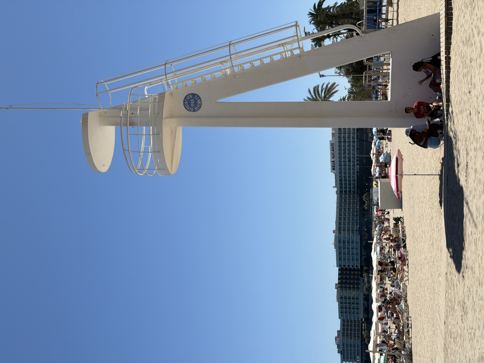 The image captures a bustling scene at Playa del Postiguet in Alicante, Spain. A lifeguard tower stands prominently, a white structure with stairs and a circular detail bearing the inscription "Diputacion Alicante 1985." Beneath the tower, individuals relax and interact. The beach is densely populated with beachgoers, umbrellas, and chairs, suggesting a peak season or a popular spot. In the background, the Melia Alicante hotel rises, providing a backdrop to the beach scene. The sky is clear and blue.