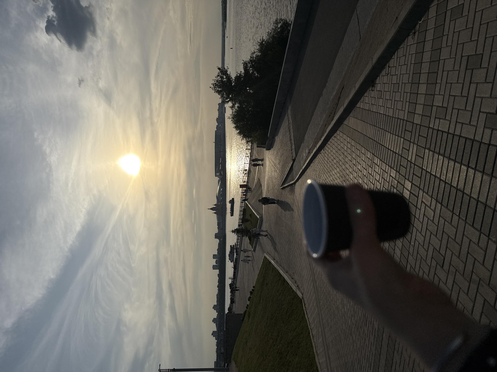 The image depicts a person holding a dark disposable cup in the foreground, with their hand and arm blurred. The scene is set on a paved walkway with a brick pattern, characteristic of the Nizhny Novgorod embankment. In the background, a wide river, likely the Volga, reflects the golden light of the setting sun. The skyline of Nizhny Novgorod is visible across the river, featuring various buildings and structures, including what appears to be a stadium. Several people are walking along the embankment, silhouetted against the warm evening light. The sky is partly cloudy with wispy cirrus clouds and the sun is low on the horizon, creating a soft, diffused light. A grassy slope with a concrete retaining wall is visible to the left of the walkway.