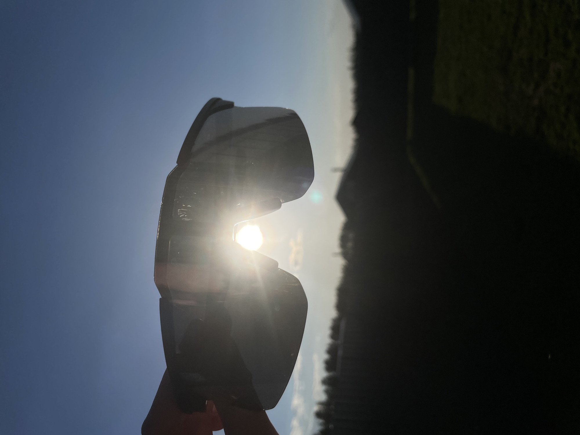 The image shows a pair of sunglasses being held up to the sun. The sun is visible through the lens. The background is blurred, but appears to be a landscape with a blue sky and some vegetation. The location might be Подшивалово, Russia, as specified.