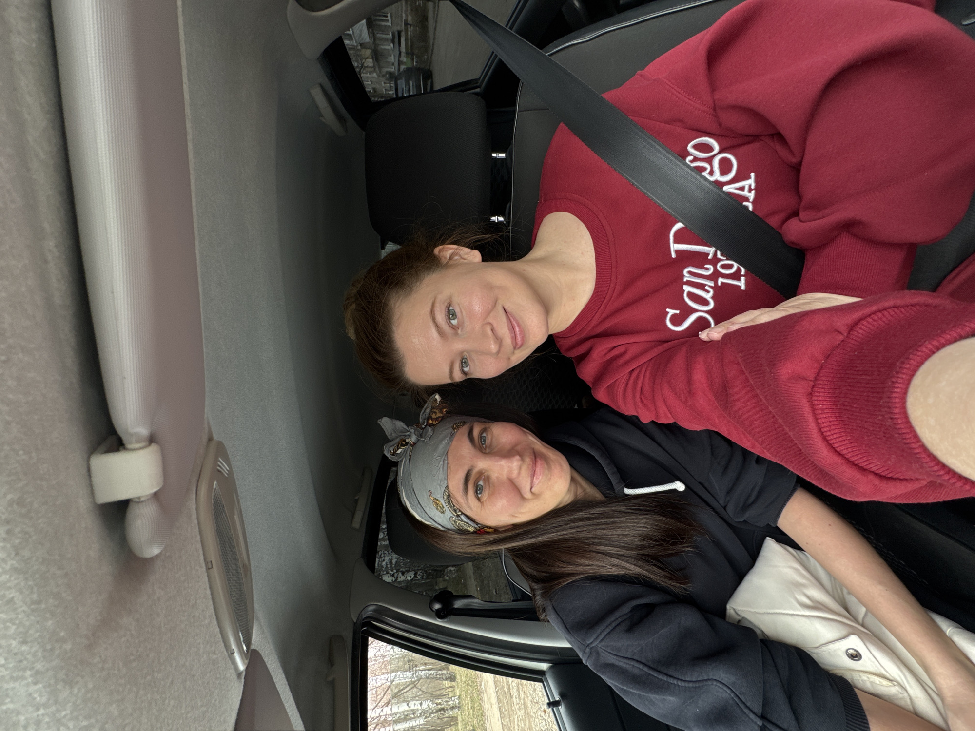 Two women are sitting in the front seats of a car. The woman on the top is wearing a red sweatshirt with white lettering. She is wearing a seatbelt. The woman on the bottom is wearing a black hoodie and a gray headscarf. The photo was taken inside of a car. Trees are visible outside the car window, suggesting the location is Chaikovsky, Russia.