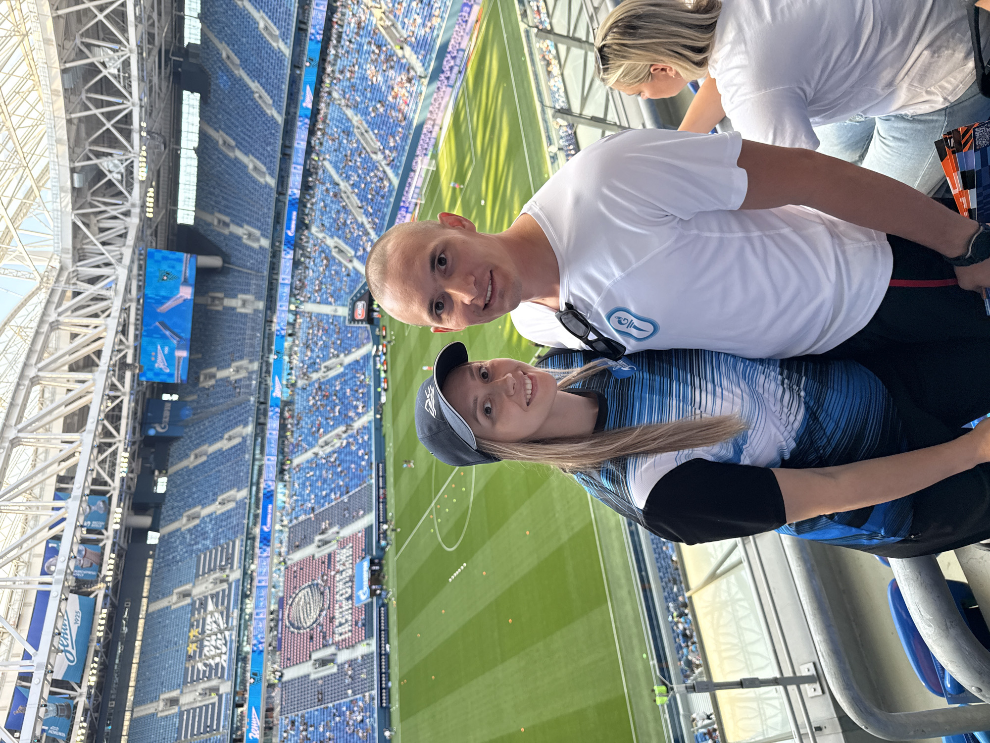 A young Caucasian couple is posing for a photo in the stands of a brightly lit, modern stadium. The stadium has rows of blue and pink seats, with a green soccer field visible below. The couple is smiling and looking at the camera. The man has a shaved head and is wearing a white t-shirt. The woman is wearing a baseball cap and a blue and black striped shirt. A portion of another woman's head and shoulder is visible on the right side of the image. The stadium is identified as Gazprom Arena in Saint Petersburg, Russia, due to the visible signage that includes "Газпром" (Gazprom) and the team's colors and logo consistent with FC Zenit Saint Petersburg. The overall atmosphere is one of enjoyment and attendance at a sporting event.