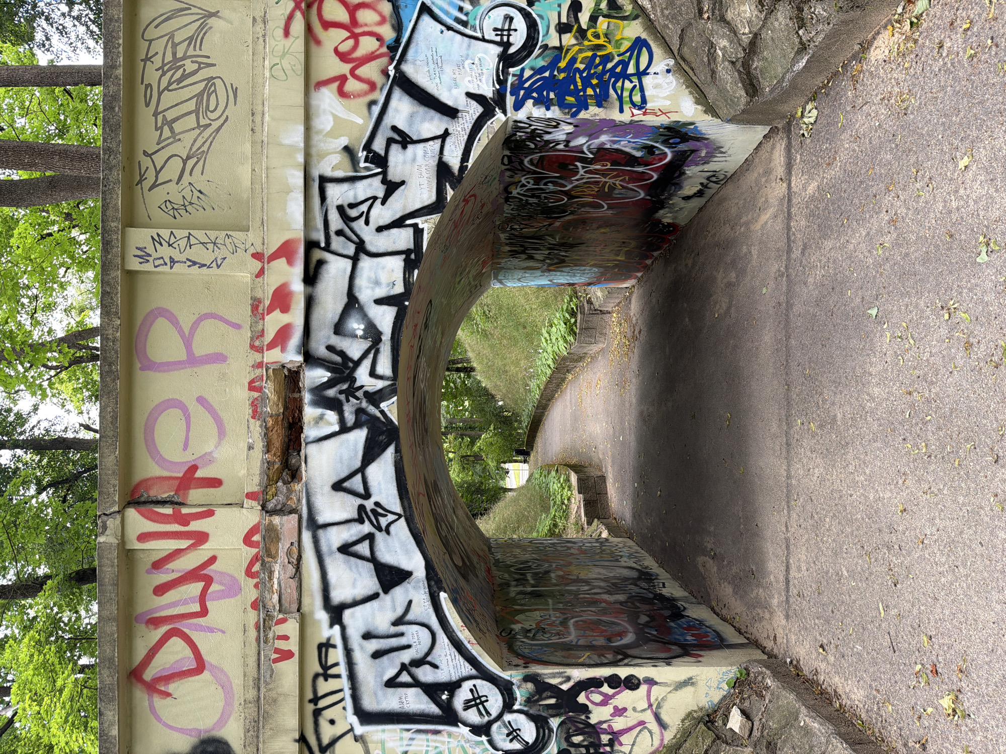 The image shows an arched underpass covered in graffiti, located in Riga, Latvia. The underpass is constructed of concrete and stone. The walls of the underpass are heavily adorned with various graffiti tags and pieces, featuring a mix of colors and styles, including red, pink, black, white, blue, and yellow. The ground inside the underpass is a paved pathway, and there are trees and foliage visible on either side of the pathway as it extends into the distance. The overall scene suggests an urban outdoor environment, likely a park or a pedestrian walkway, that has been subjected to graffiti.