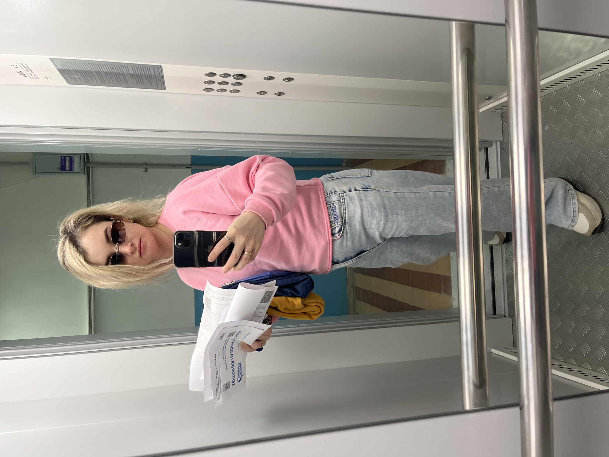 A woman with blonde hair and sunglasses is taking a mirror selfie in what appears to be an elevator or small lift. She is wearing a pink sweater and light-wash jeans, holding a smartphone in her right hand and several papers in her left hand. Behind her are elevator controls and a metal handrail. The elevator floor is made of textured metal, and there is a glimpse of a building outside the elevator window. The papers she is holding appear to be documents. There are clothes hanging from the inside of the shirt. The location is not specifically provided but, based on the user's prompt, would be in the city of Chaikovsky, Russia.