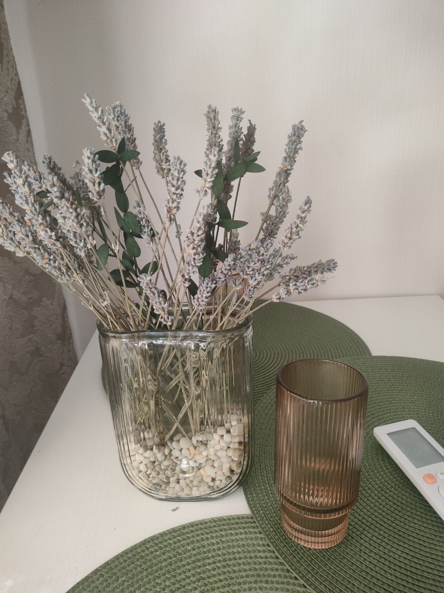 This is a still life photograph taken indoors, likely in a domestic setting. The image features a bouquet of dried lavender in a clear, ribbed glass vase filled with small, light-colored pebbles. To the right of the vase, on a textured green placemat, sits a tall, ribbed amber glass. A white remote control is also present on the green placemat, positioned to the right of the amber glass. The background consists of a plain, light-colored wall and a draped curtain on the left. The lighting suggests an indoor environment, possibly during the daytime. There are no people or animals visible in the image. There is no information in the image to suggest its location in Lyubertsy, Russia.