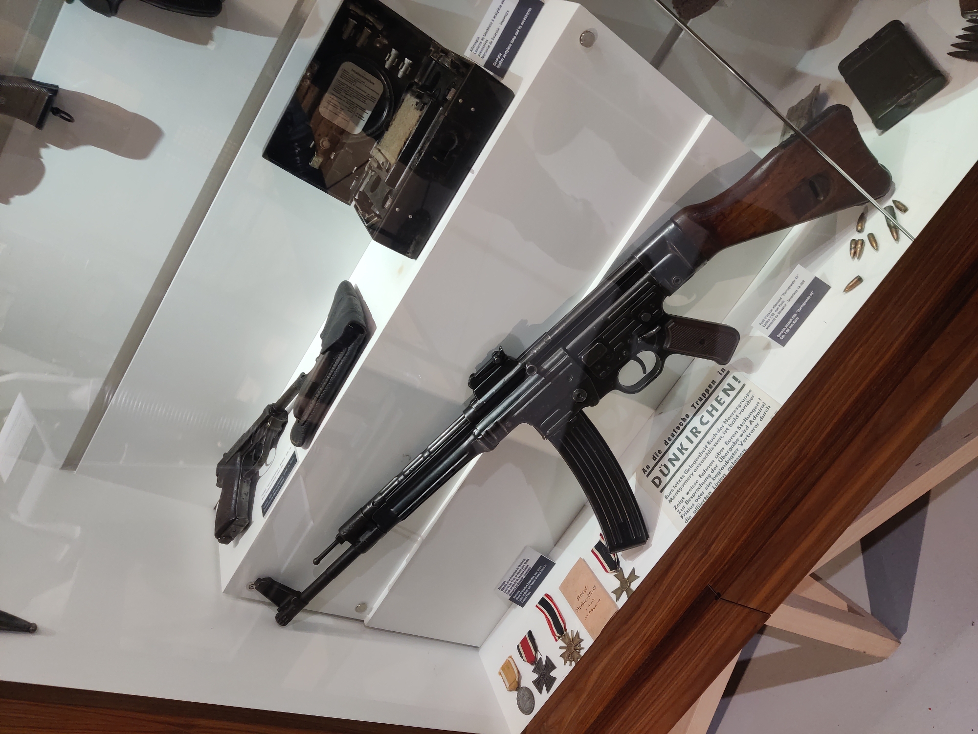 The image shows a museum display case containing military artifacts. The primary object of focus is a German StG 44 assault rifle, complete with a wooden stock and a ribbed magazine. To the left of the rifle, there are several medals with ribbons, including what appears to be an Iron Cross. A small document with text is also present near the medals. Above and to the right of the rifle, a black case is open, revealing what appears to be a lighting or signaling device with accessories. Further in the background, a pistol is visible on a shelf. Below the rifle, several bullets are scattered on the shelf, and a small green rectangular object, possibly an ammunition box or accessory case, is present. A sign with German text, mentioning "DÜNKIRCHEN!", is also visible. The display is set against a white background and is enclosed in a glass case, with wooden framing visible at the bottom and left. The overall scene is a static museum exhibit of historical military items, with no people or actions depicted. The presence of the "DÜNKIRCHEN!" text on the sign suggests the display relates to events in Dunkirk, France.
