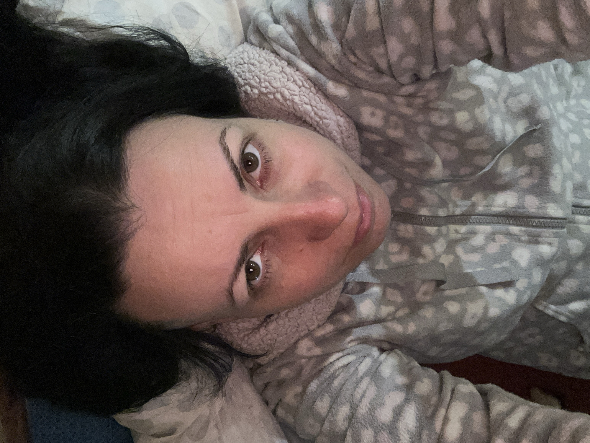 A woman with dark hair lies on a bed in a gray and white patterned hooded sweatshirt. Her face is close to the camera, tilted upward. The background shows a bed with patterned sheets and part of a wooden headboard or frame. No information about the location in Sofia, Bulgaria, is discernible from the image.