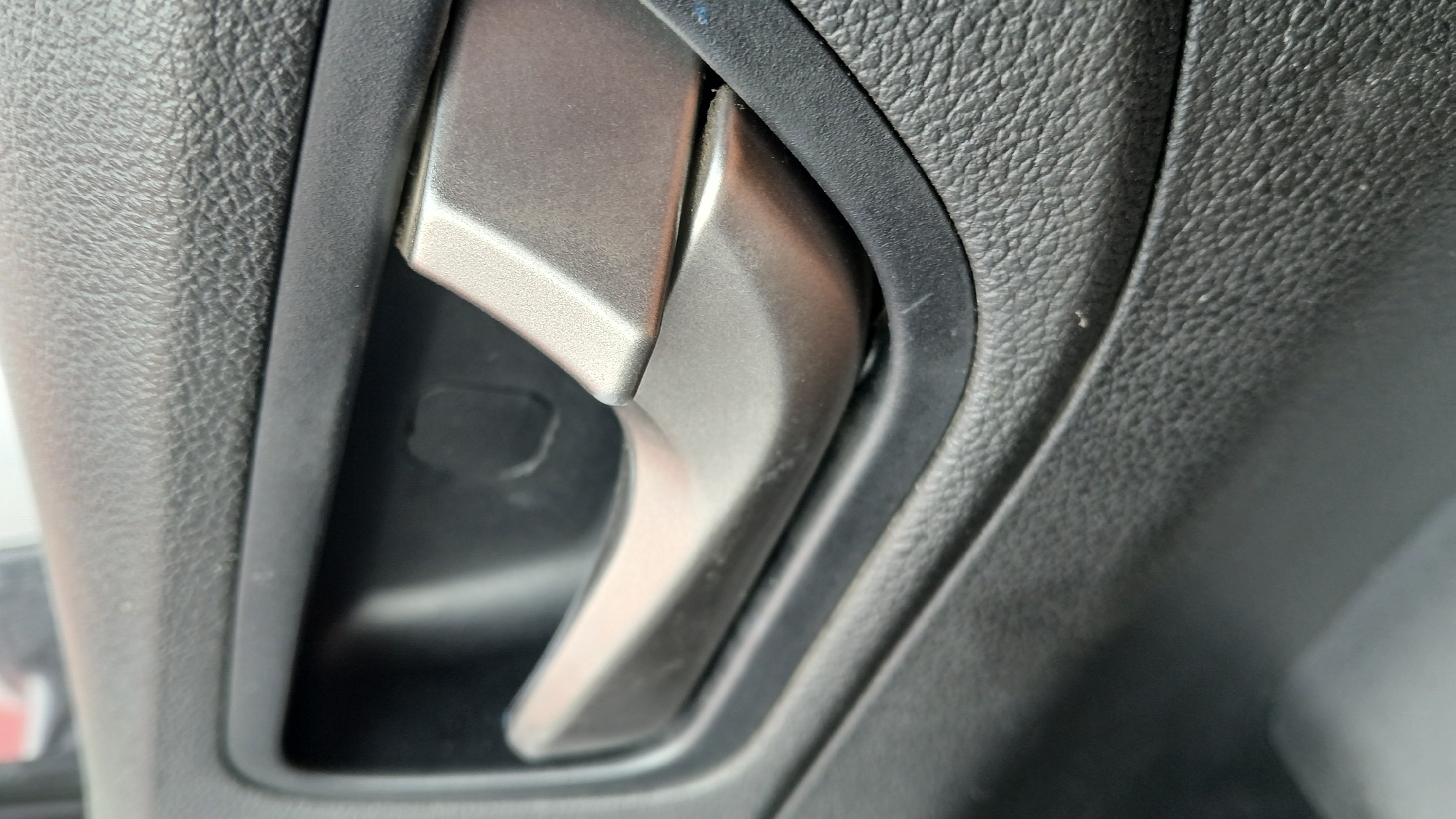 The image shows a close-up of a car's interior door handle. The handle is made of a metallic gray material and is shaped to be gripped. It is set into a textured black plastic panel, likely the door card of the vehicle. The background is blurred but suggests a car interior environment. There are no people visible in the image. The image does not provide any information about the specific location within the city of Rubtsovsk, Russia.
