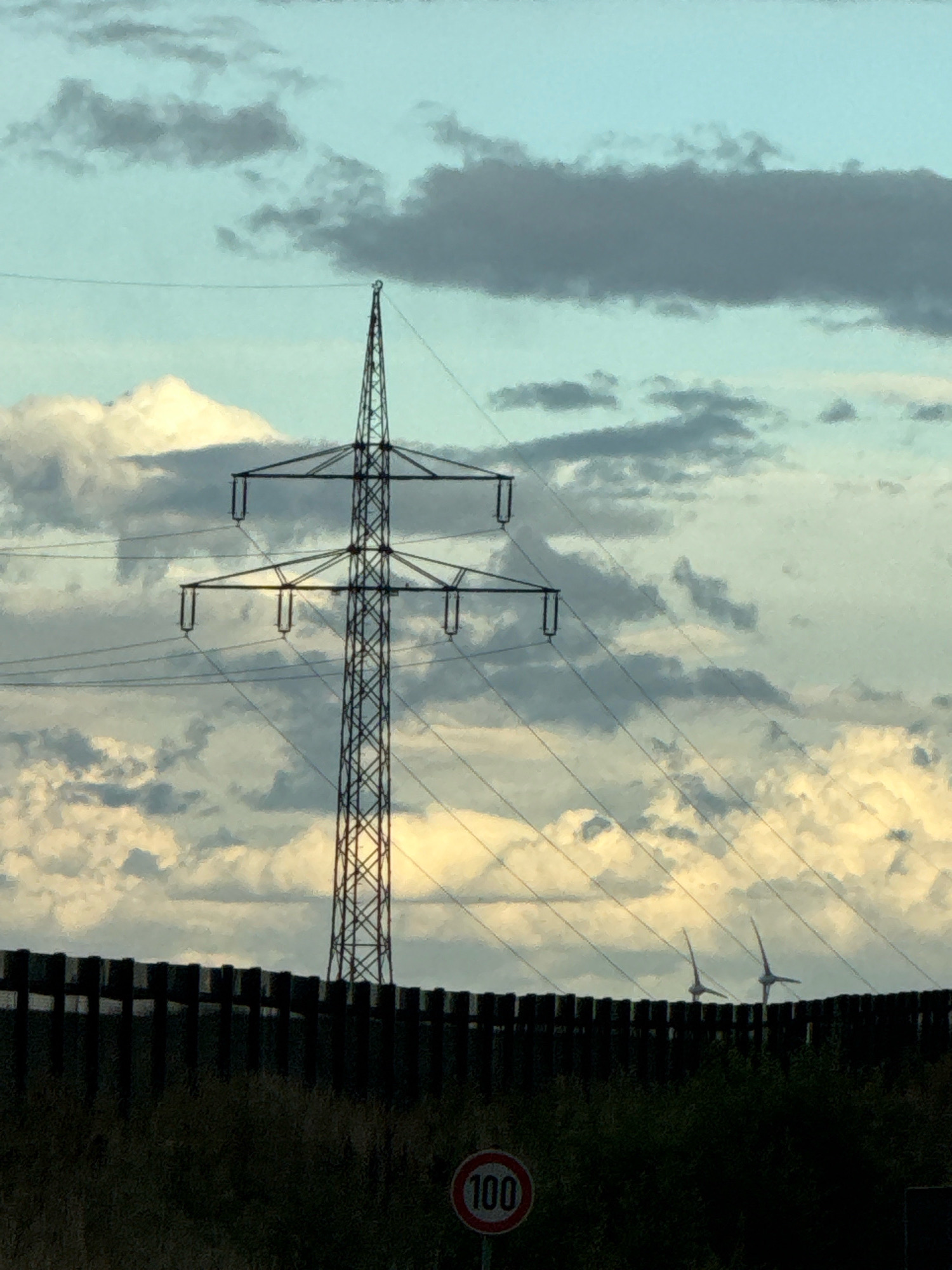 An outdoor, low-angle shot captures a metal electricity pylon against a cloudy sky with a hint of sunset. The pylon is tall and lattice-like, with multiple crossbars supporting thick cables. A dark wooden fence runs horizontally across the foreground, partially obscuring lush green vegetation. In the distance, beyond the fence, two wind turbines are visible, their blades turned towards the left. A circular traffic sign with "100" in red on a white background is positioned near the bottom right of the frame. The scene is likely located in Bad Lauterberg im Harz, Germany, given the presence of wind turbines, a common feature in the region. The lighting suggests it's either early morning or late afternoon.