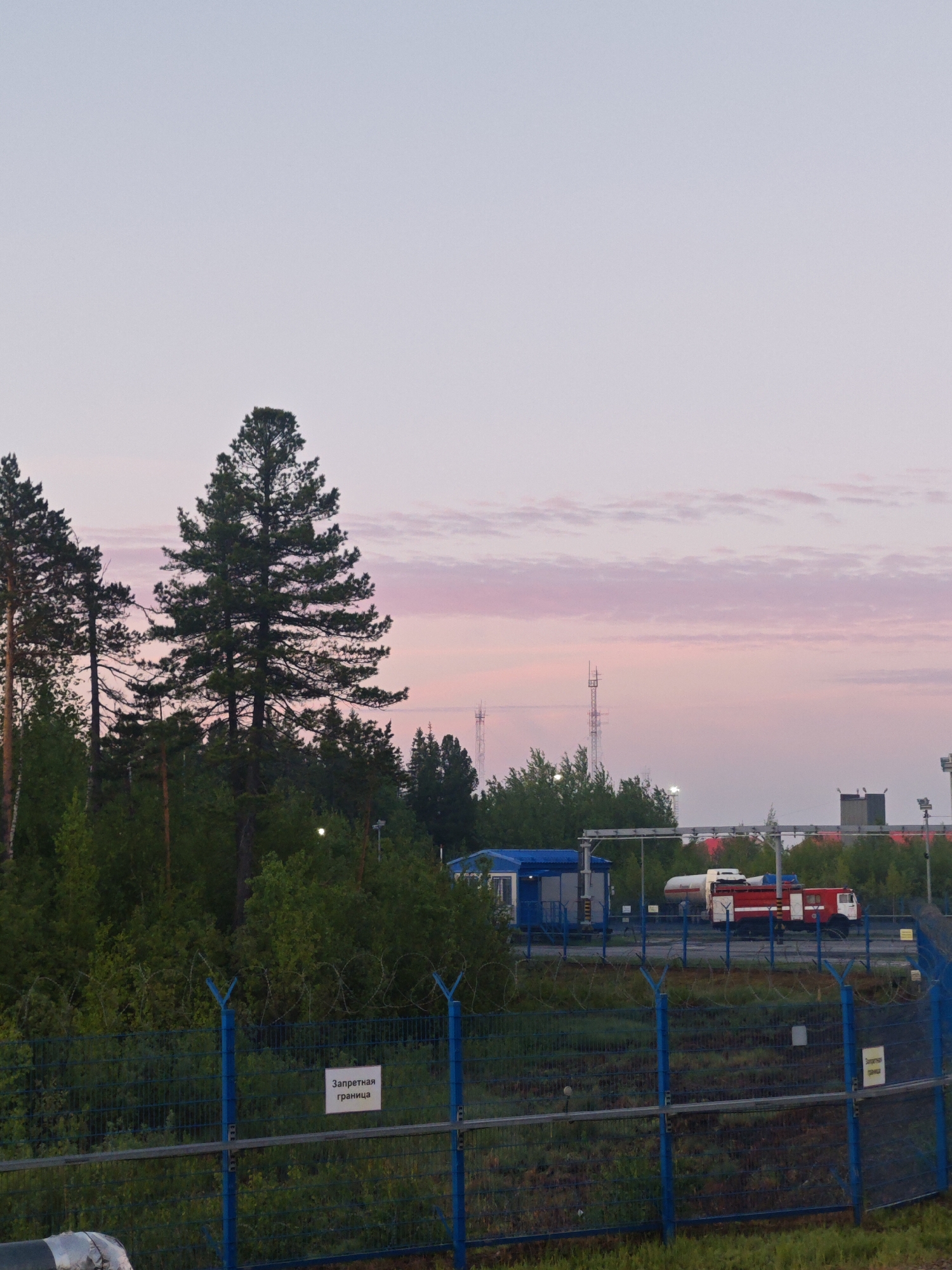 The image depicts an outdoor scene at either dawn or dusk, characterized by a pastel sky displaying shades of light blue and soft pink, with faint, wispy clouds. The foreground and midground are dominated by a blue metal fence topped with barbed wire, clearly marking a boundary.
Behind this fence, a lush, dense forest of tall evergreen trees, likely pines, occupies the left side of the frame. To the right of the forest, within the fenced area, a blue modular building or container is visible, along with industrial structures and pipes. A red and white tank truck, resembling a fuel or fire truck, is parked further back on a paved surface. In the distance, several tall, slender metal towers or masts rise above the treeline, silhouetted against the sky. A large, insulated pipe is partially visible in the bottom left foreground.
No people are present in the scene, suggesting a calm and operational environment. A white sign attached to the blue fence displays the Russian text "Запретная граница" (Zapretnaya granitsa), which translates to "Forbidden boundary" or "Restricted area," indicating controlled access to the site. The overall setting combines natural forest elements with industrial infrastructure, typical of remote or resource-focused areas in Russia. The soft lighting creates a tranquil, somewhat isolated atmosphere.