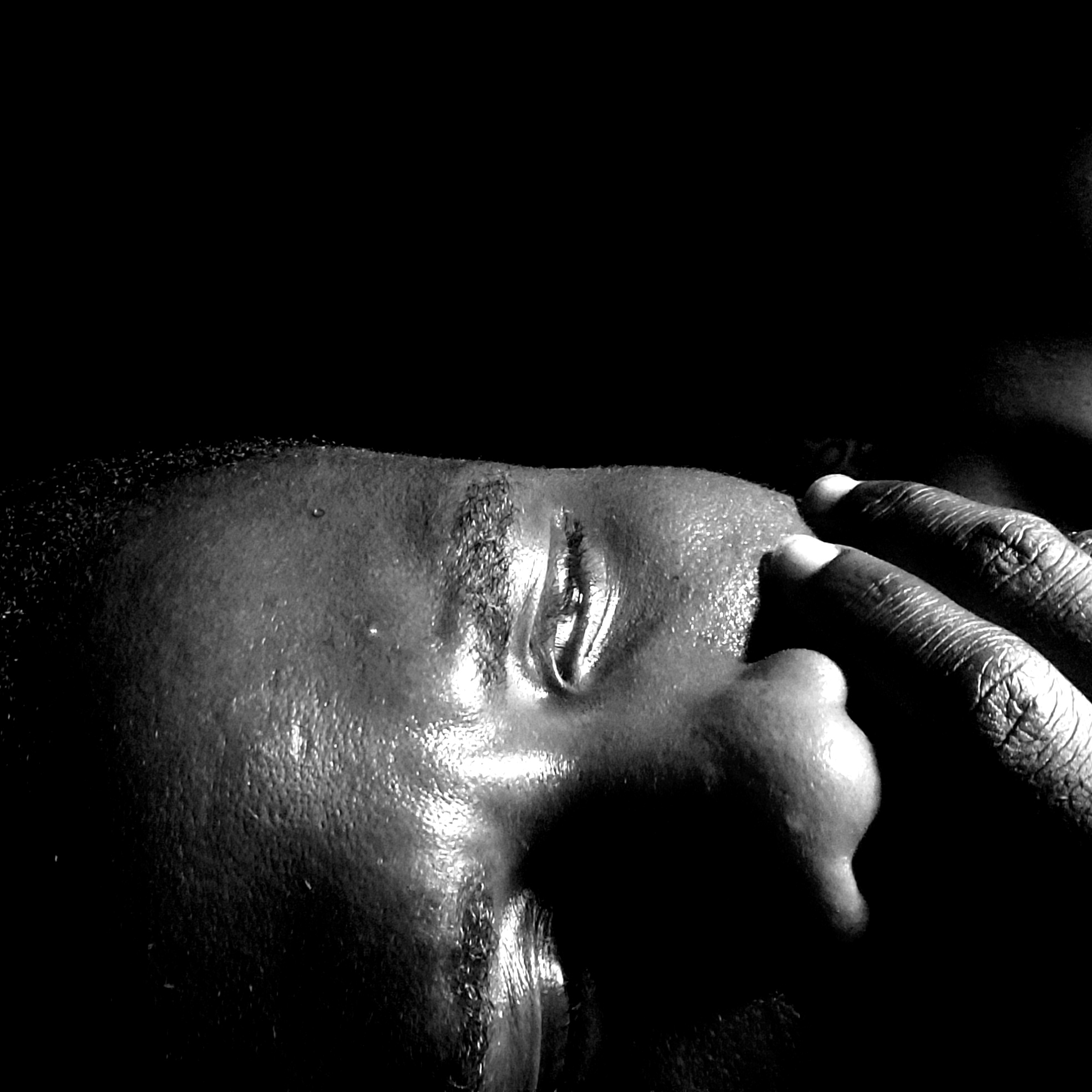 A close-up, black and white photograph depicts a person's face in low lighting conditions. The individual has dark skin and close-cropped hair. A single eye is visible, with the face oriented at an angle. The person's fingers are pressed against their face. The scene's location in Narayi, Nigeria, cannot be determined from the provided visual details.