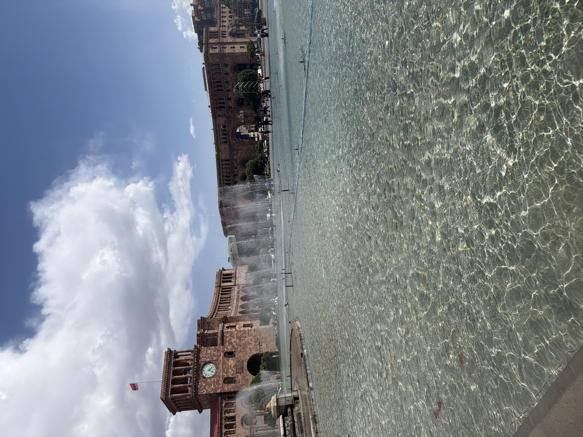 The image depicts a partially obscured view of Republic Square in Yerevan, Armenia, featuring the Singing Fountains. The fountains occupy a significant portion of the frame, with water jets visible. To the left are buildings constructed of dark brown stone, including a clock tower displaying a working analog clock and the Armenian flag. People are partially visible near the buildings. The sky is partially obscured by the buildings, and shows patchy clouds.