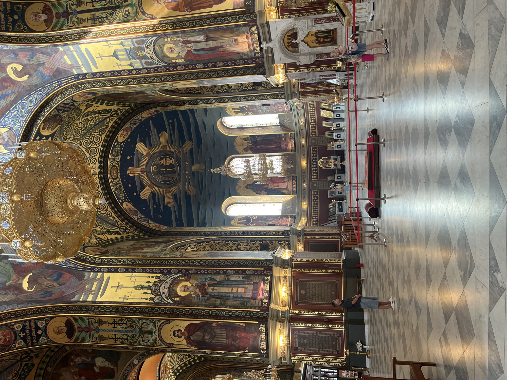 The image shows the interior of the Ascension Cathedral in Novocherkassk, Russia. The cathedral is ornately decorated with gold accents, frescoes, and large chandeliers. In the foreground, a man wearing a dark t-shirt and denim shorts stands facing away from the camera, looking towards the altar area. To the right, a woman with blonde hair in a striped t-shirt and denim skirt is standing with a young child wearing a pink dress. They are looking towards the altar. Red velvet ropes and stanchions are set up to guide visitors. The floor is tiled in a diamond pattern of white and gray marble. The walls and ceiling are covered in religious artwork and gold detailing.