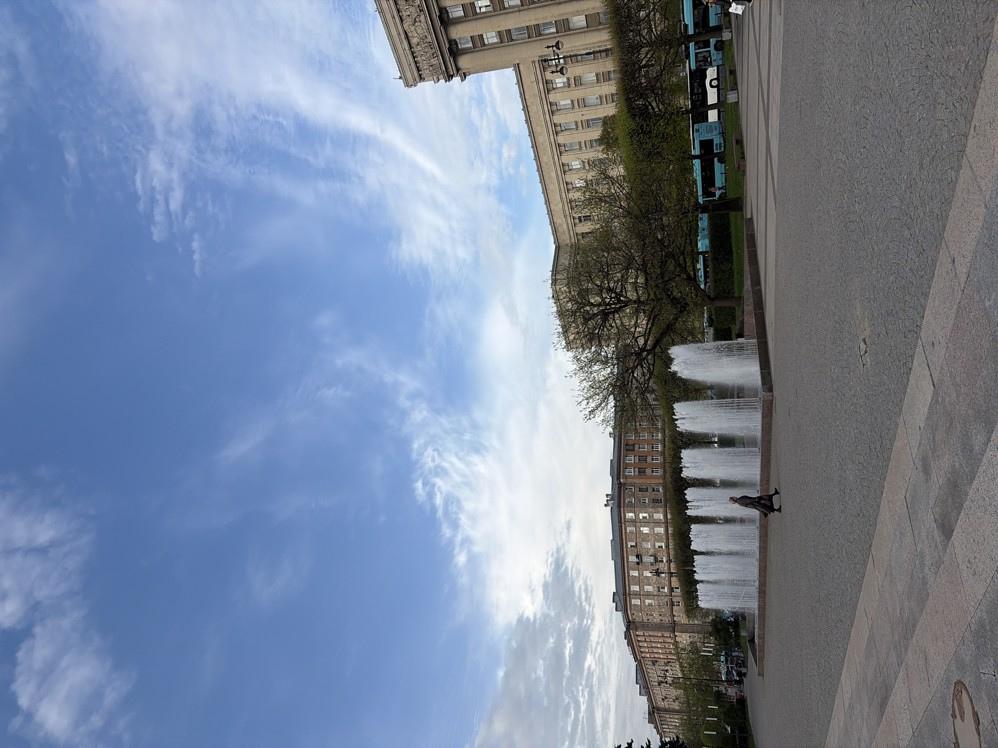 The image shows a street scene in Saint Petersburg, Russia. The perspective is angled, featuring buildings on either side and a blue sky with wispy clouds. A series of fountains is visible on the right, with a person walking near them. On the right, a building, trees and buses are also observed. The scene is captured in daylight.