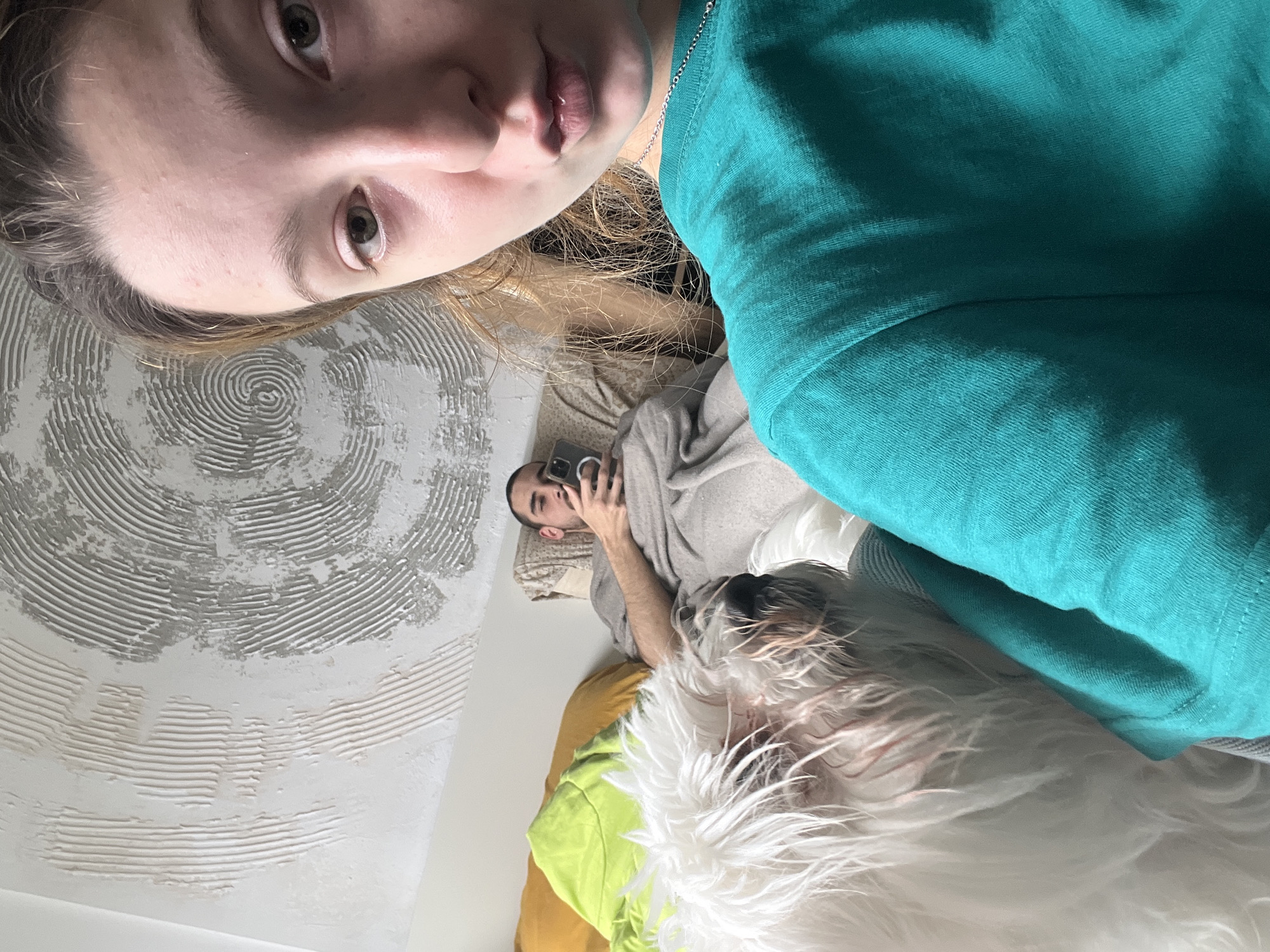 The image depicts three individuals and a dog in an indoor setting.
A person, presumably taking the photo, is positioned at the top of the frame, displaying their face and upper body. They have light skin, light brown hair, and are wearing a turquoise shirt. A silver necklace is visible around their neck. The background behind them includes a wall with a textured, swirling pattern.
Below this person, a second individual lies on a couch or bed, holding a mobile phone as if taking a photo or video. This person has short dark hair and is wearing a light gray shirt. The person is lying on light colored pillows.
The third individual is a long-haired, white dog.
The environment includes cushions in yellow and green. The location is unidentifiable, but the request indicates that it may be in Varna, Bulgaria. No specific landmarks or features are discernable to confirm this location.