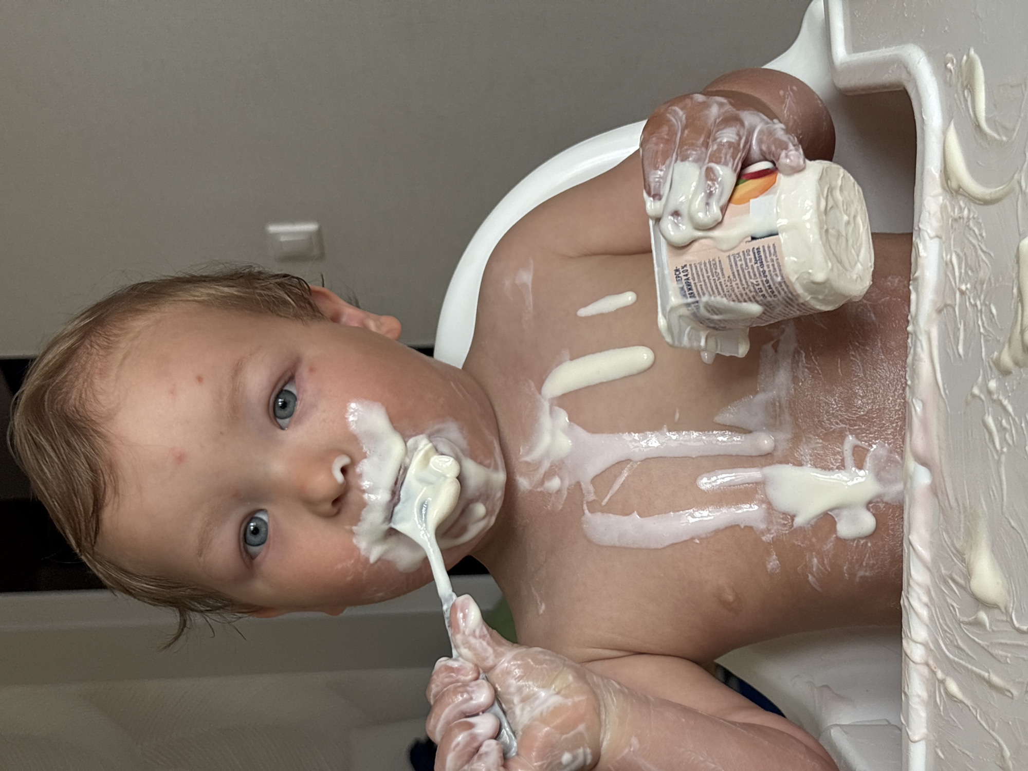 A baby is sitting in a high chair, covered in yogurt. The baby is holding a spoon in one hand and a yogurt container in the other, both covered in yogurt. Yogurt is smeared on the baby's face, chest, and the high chair tray. The baby has blue eyes and light hair. The location is not discernible from the image. I cannot determine if this image was taken in Chaikovsky, Russia.