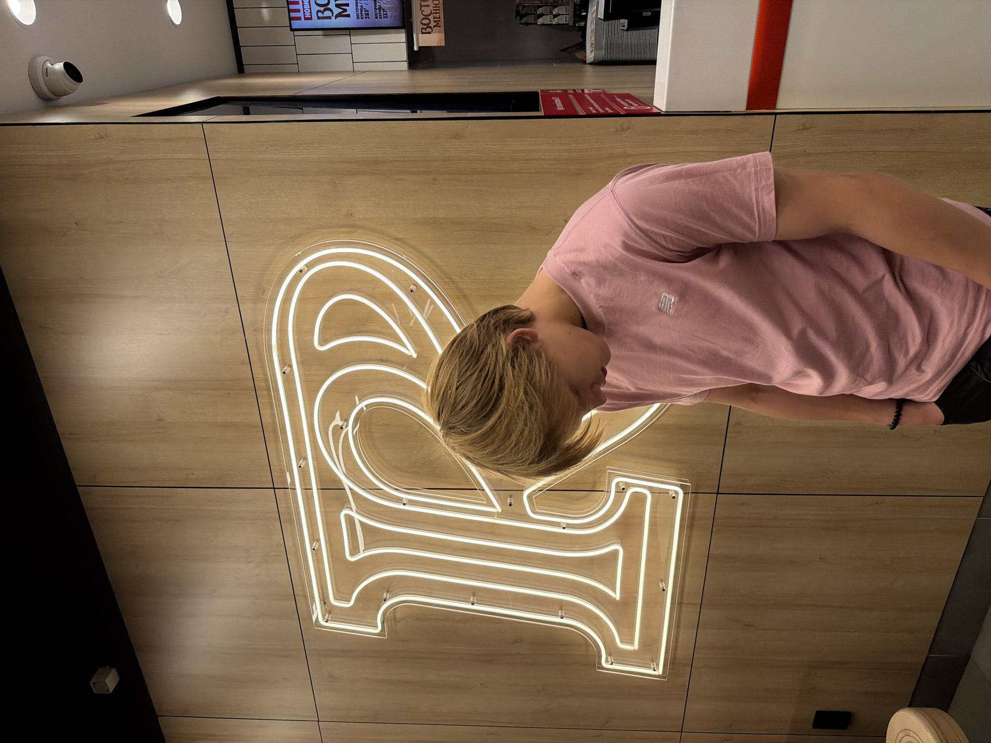 A young person with blond hair is standing in front of a wall with a large neon sign shaped like the letter "R". The sign is white and outlined in neon tubing. The wall is made of wood-paneled panels. The person is wearing a pink t-shirt with a logo on the left chest and black pants. They are looking down, with their head bowed. A surveillance camera is visible on the ceiling in the upper right corner. Part of a menu board is visible on the right side of the image. The overall setting appears to be an indoor establishment, possibly a restaurant or cafe, in Izhevsk, Russia.
