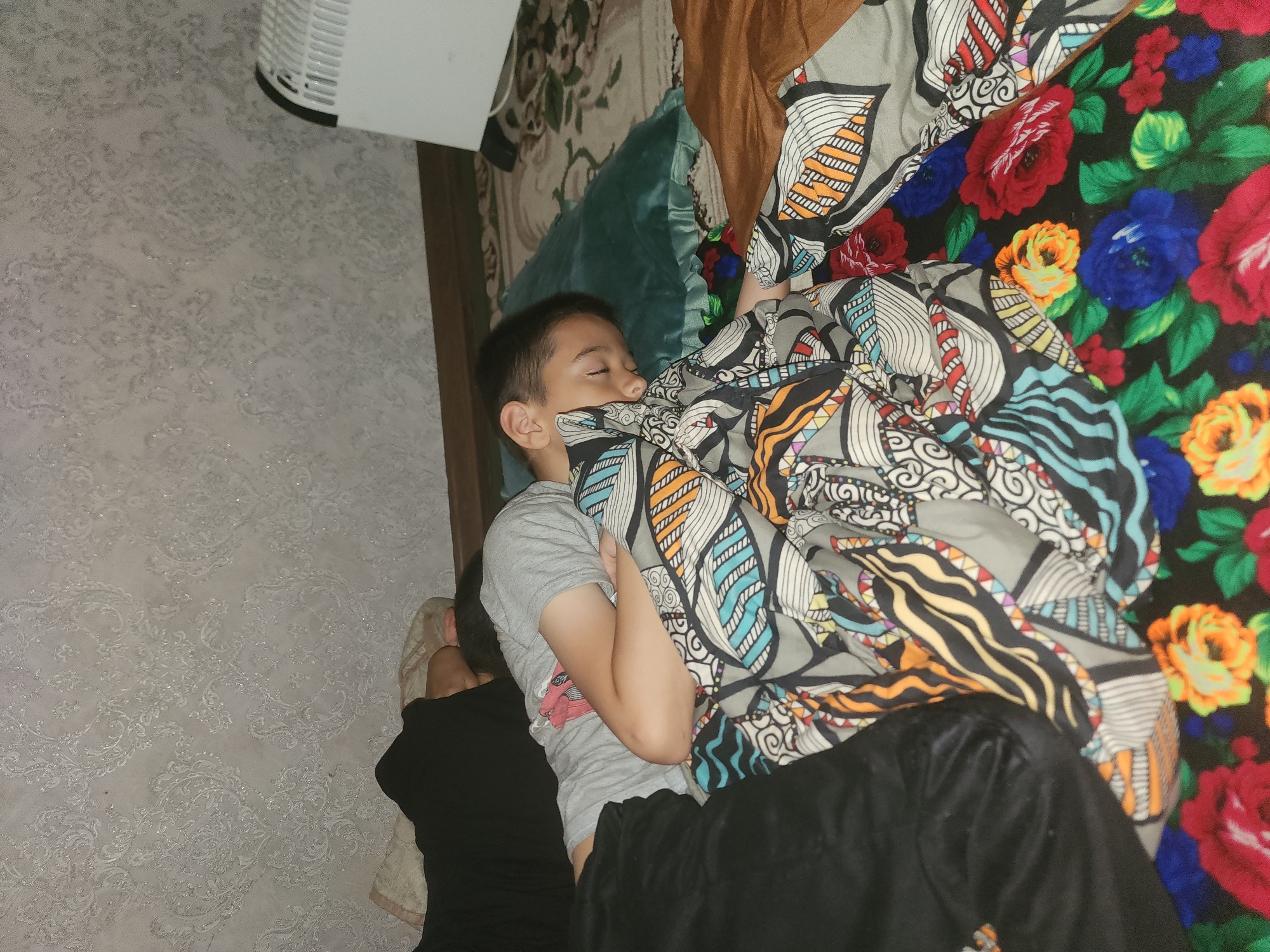 A young boy sleeps on a bed, his head resting on a teal pillow. He is covered by a blanket with a colorful floral pattern, featuring red, blue, yellow, and green flowers. Another blanket with a geometric print in shades of gray, blue, and orange is also visible. The boy is wearing a light gray t-shirt. To the left, a person wearing black clothing is partially visible, also appearing to be resting. The background shows a wall with patterned wallpaper and a white radiator on the right side of the image. The scene appears to be indoors, likely a bedroom in a residential setting in the city of Yefremov, Russia.