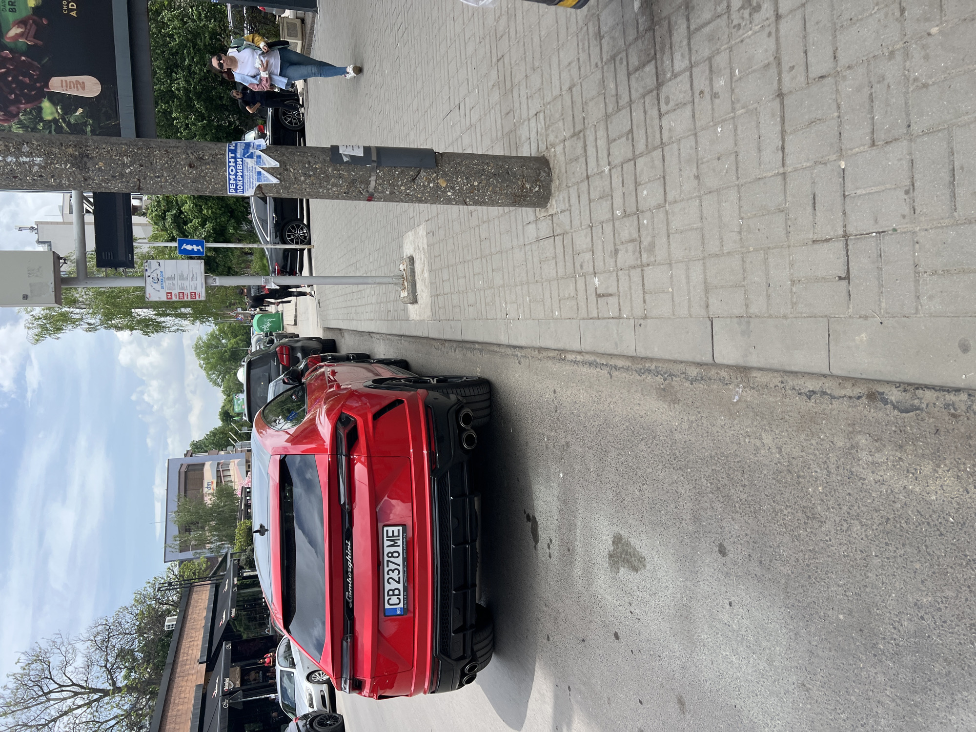 The image depicts a red Lamborghini Urus parked on a sidewalk in Sofia, Bulgaria. The license plate number is CB 2378 ME. In the background, a person is walking along the sidewalk, while other cars are parked along the street. A utility pole is on the sidewalk, and a bus stop sign and a poster with "РЕМОНТ HA ПОКРИВИ" are attached to it. There's an ad for Nuii ice cream in the background. A building or establishment with a black awning is visible near the red car. The sidewalk is paved with grey bricks.