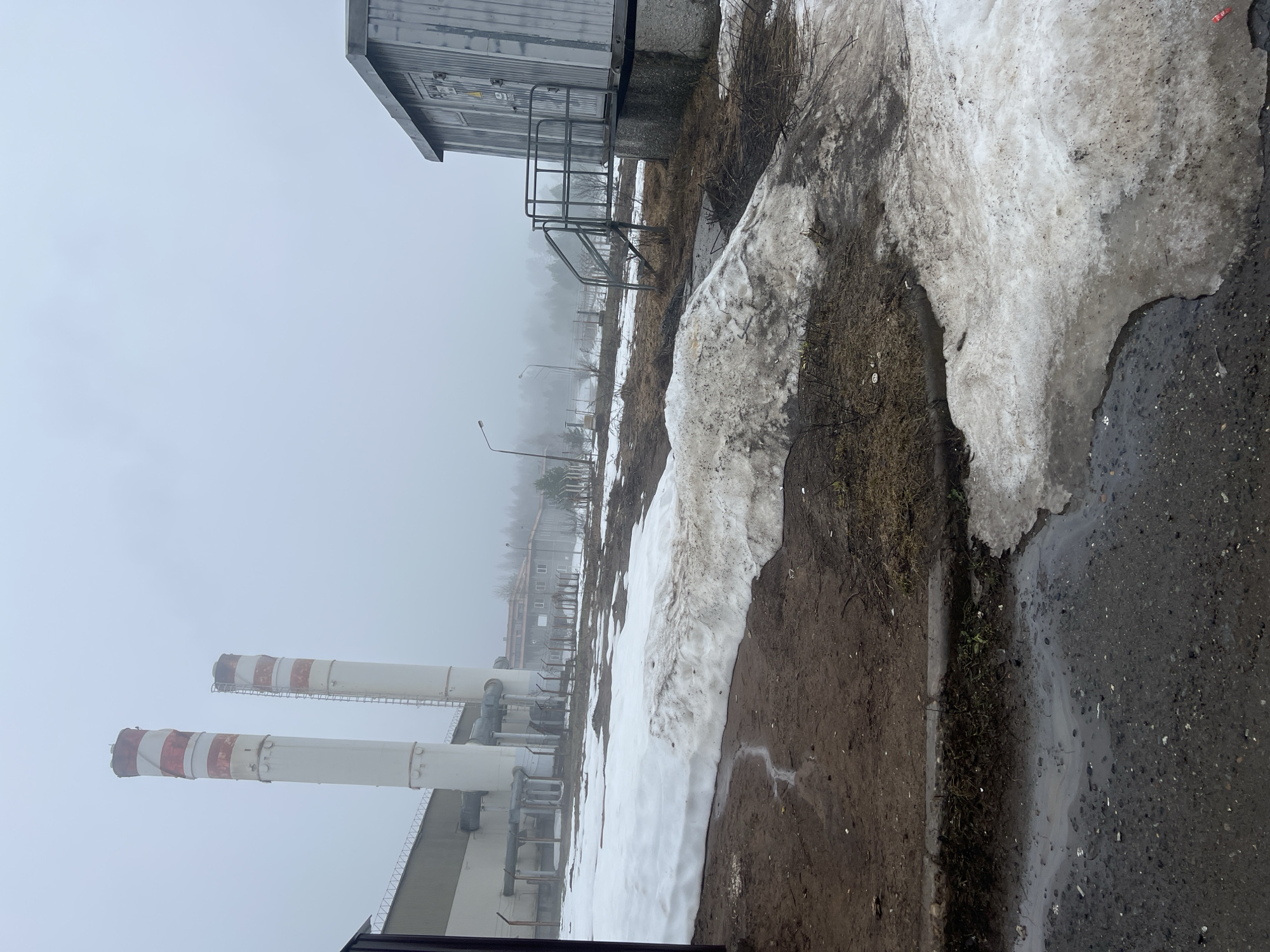 The image depicts an industrial area in Марковский, Russia on a foggy day. Two tall, white smokestacks with red and white striped tops are visible in the distance. The smokestacks are part of a larger industrial building. In the foreground, there is a patch of melting snow along a paved area. A small metal building with stairs is also visible. The ground is muddy and wet. The overall scene suggests a cold, industrial environment.