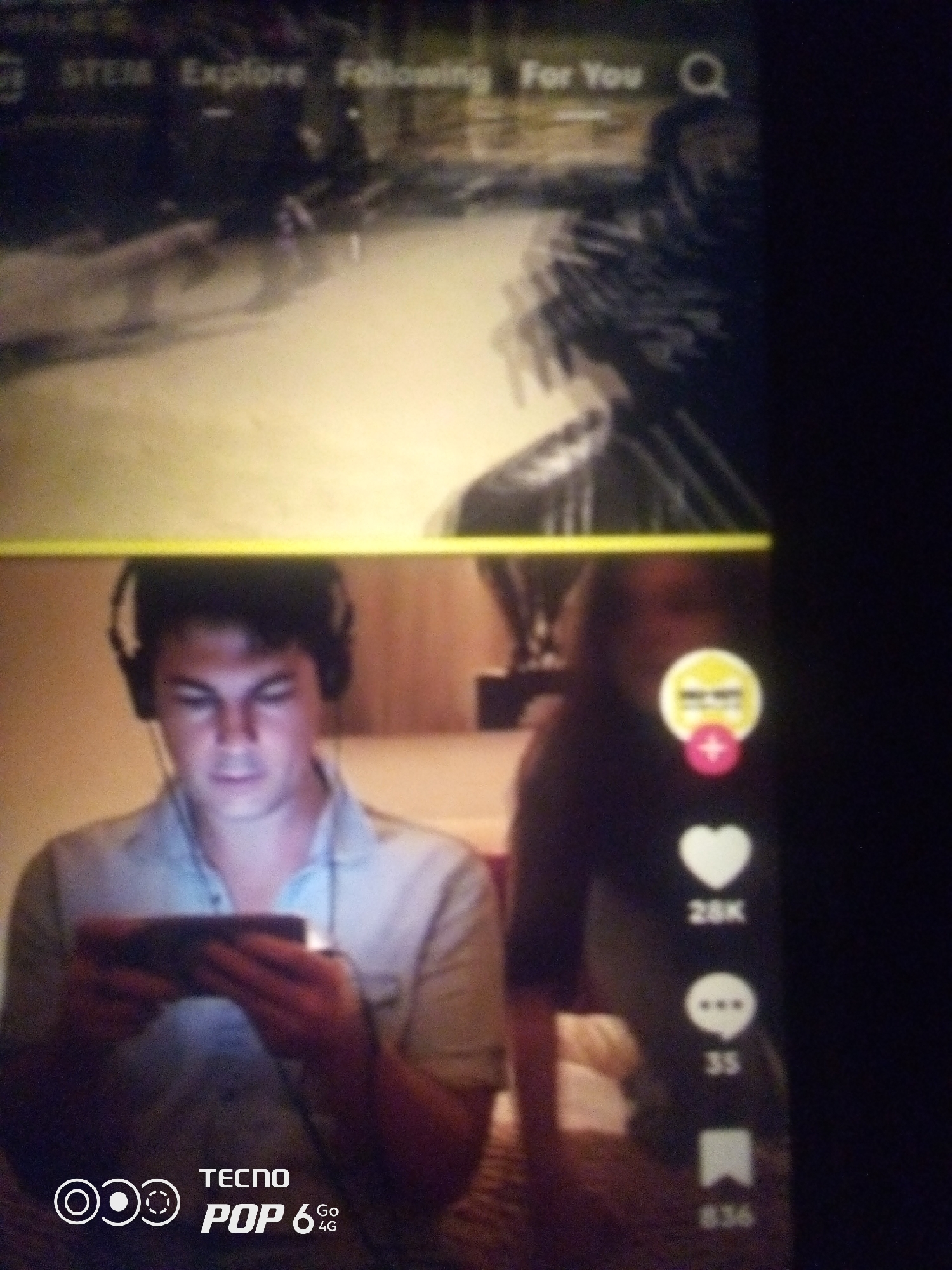 The image is a screenshot of a mobile device displaying a social media application, likely TikTok. The top portion of the screen shows a video with a blurry background that appears to be an indoor scene. Text elements like "STEM," "Explore," "Following," and "For You" are visible, along with a search icon.
The bottom portion of the screen features a young man wearing headphones and looking down at his phone, which he is holding in his hands. He is wearing a light-colored shirt. To the right of the man, on the social media interface, there are icons indicating likes, comments, and shares, with corresponding numbers (28K likes, 35 comments, 836 shares). A circular icon with a pink plus sign is also visible.
At the very bottom of the screen, there is a logo that reads "TECNO POP 6 4G."
The image does not provide any information to determine the specific location within the city of Howayi, Nigeria, nor does it offer details about the context or purpose of the video being viewed. It is a digital capture of a user interacting with a mobile application.