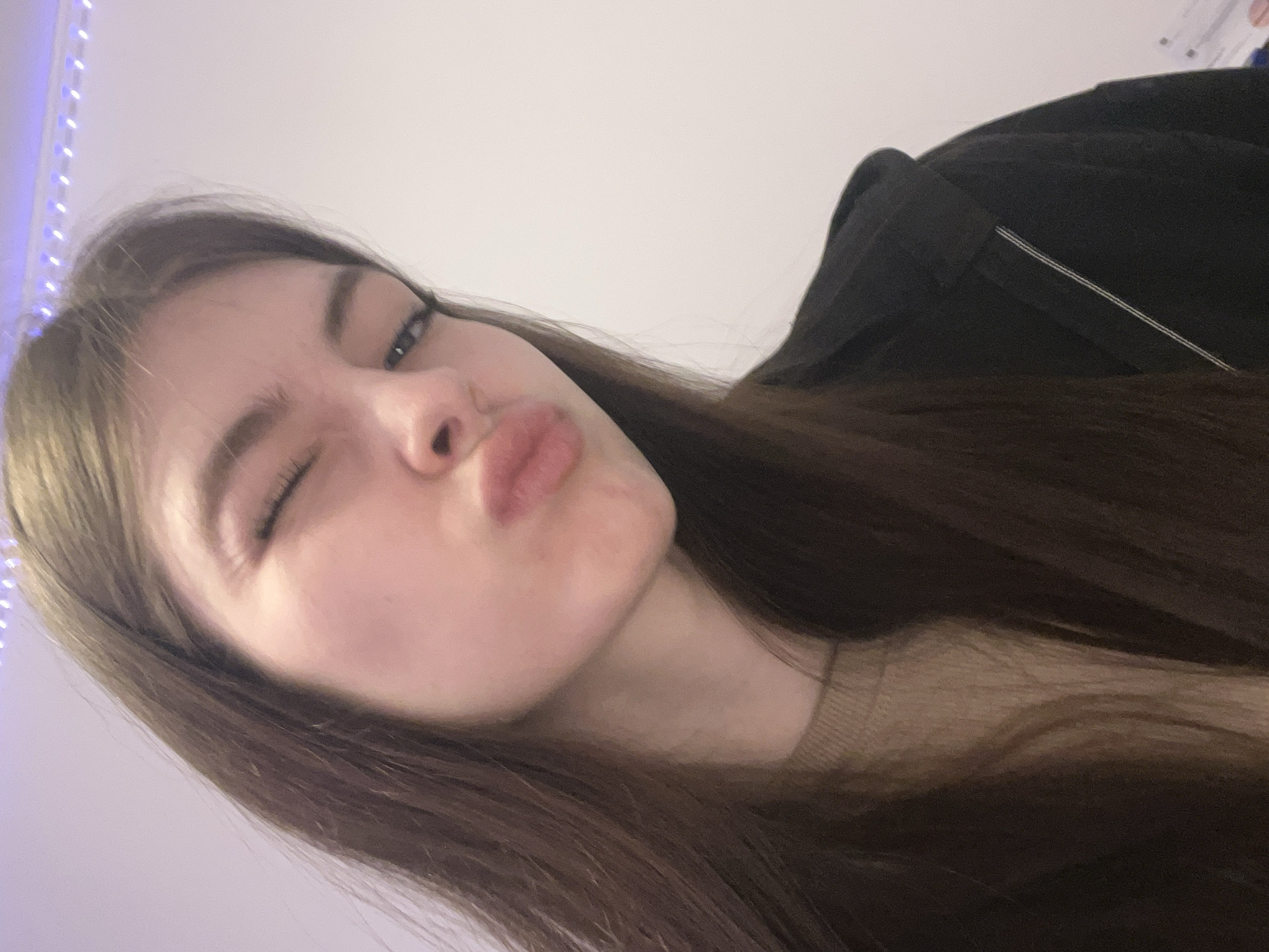 A young woman with light skin, long brown hair, and brown eyes is posing for a selfie. Her lips are pursed in a kissy face and one eye is closed in a wink. She is wearing a light-colored sweater and a dark jacket. A string of lights is visible in the upper left corner of the frame. The background is a plain white wall. The location is not discernible as Saint Petersburg, Russia.