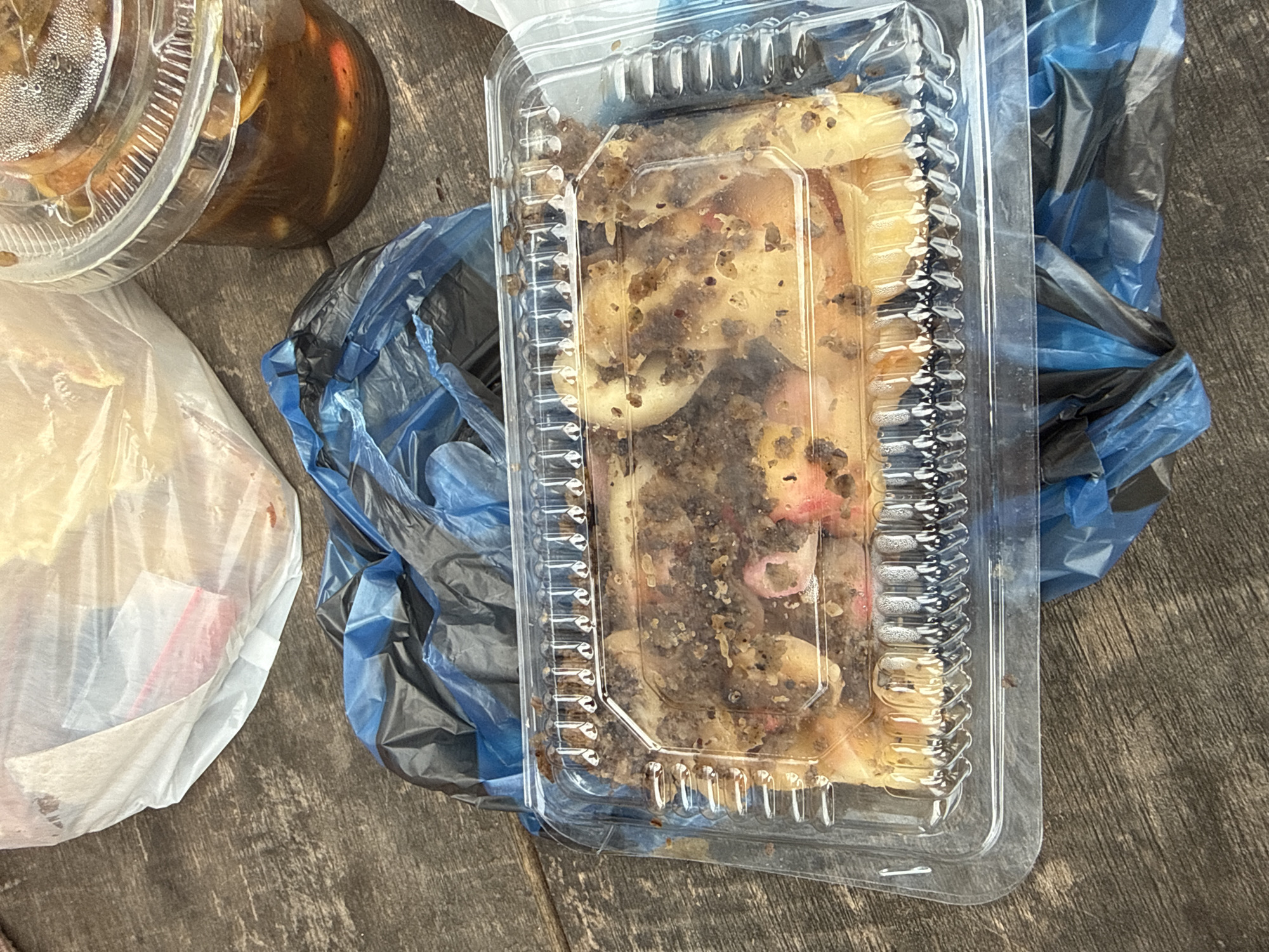 The image shows food items and plastic bags on a wooden surface. In the foreground, a clear plastic container holds a portion of food, possibly a dessert or pastry, with a granular, dark topping. The food appears to be light-colored with some reddish-pink elements, and it is covered in crumbs or small pieces. The container has a ridged lid. To the right of the container is a black and blue plastic bag, with the blue part of the bag appearing more prominently. To the left of the container, there is a white plastic bag containing some items, including what looks like a white paper napkin and a small red and white packet. Further to the top left, another clear plastic container is partially visible, containing a dark liquid and some solid pieces. The overall scene suggests a discarded meal or leftover food items. The wooden surface has a weathered texture, indicating an outdoor or rustic setting. No people or specific text are visible in the image. The lighting suggests it might be daytime, but no strong shadows indicate a specific time. The location context provided is Meunasahbale, Indonesia.