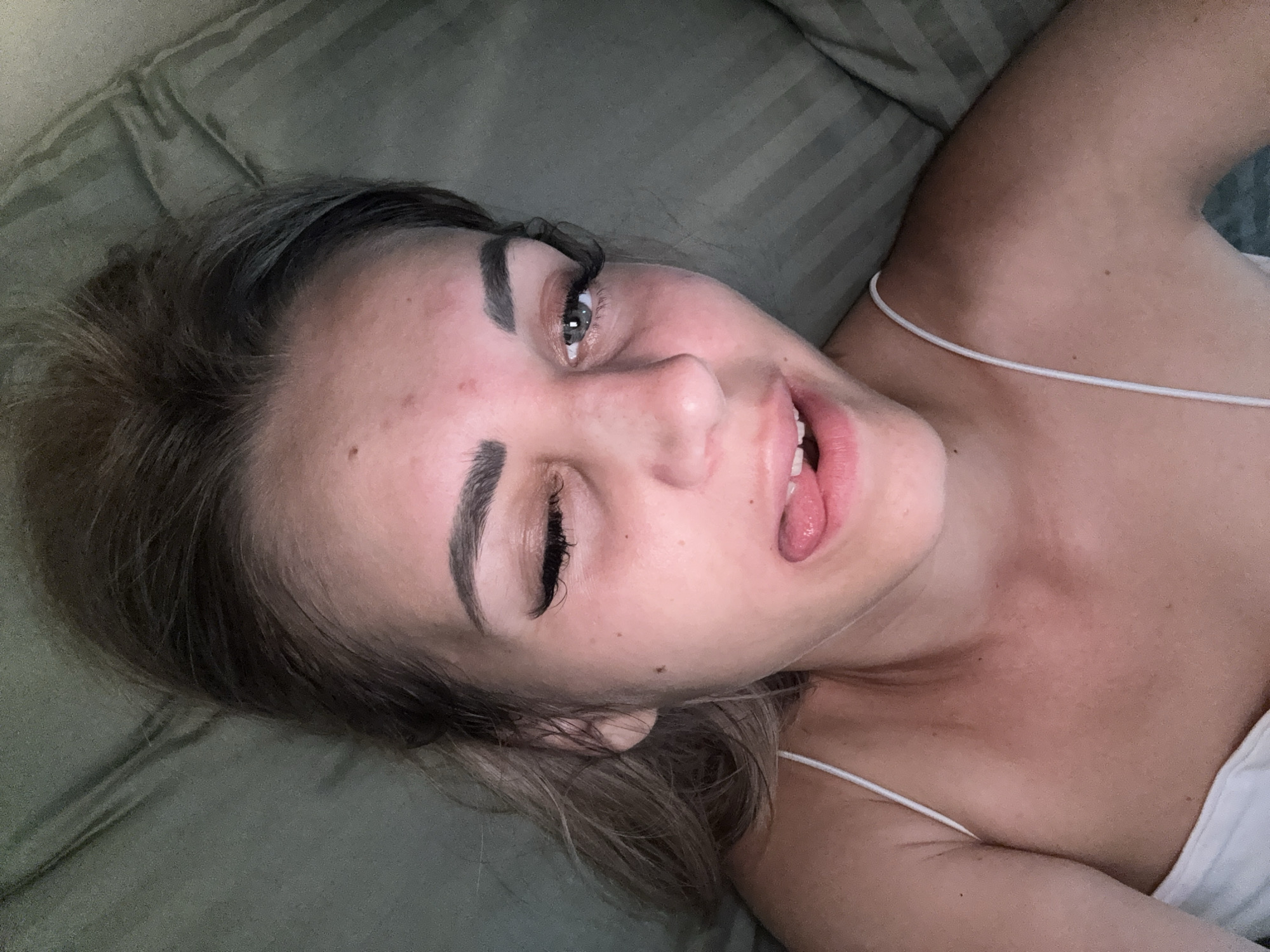 The image shows a close-up, overhead view of a young woman lying on a textured, greenish-gray surface, possibly a couch or bed. She has light brown hair and is wearing a white tank top with thin straps. Her head is tilted, and she is looking towards the camera with one eye open and the other partially closed, her tongue sticking out slightly. Her makeup includes prominent eyelashes and defined eyebrows. The background consists of the textured surface, which appears to be fabric with a striped pattern. There are no discernible features or landmarks that would indicate a specific location in Yuzhno-Sakhalinsk, Russia. The overall scene is intimate and personal.