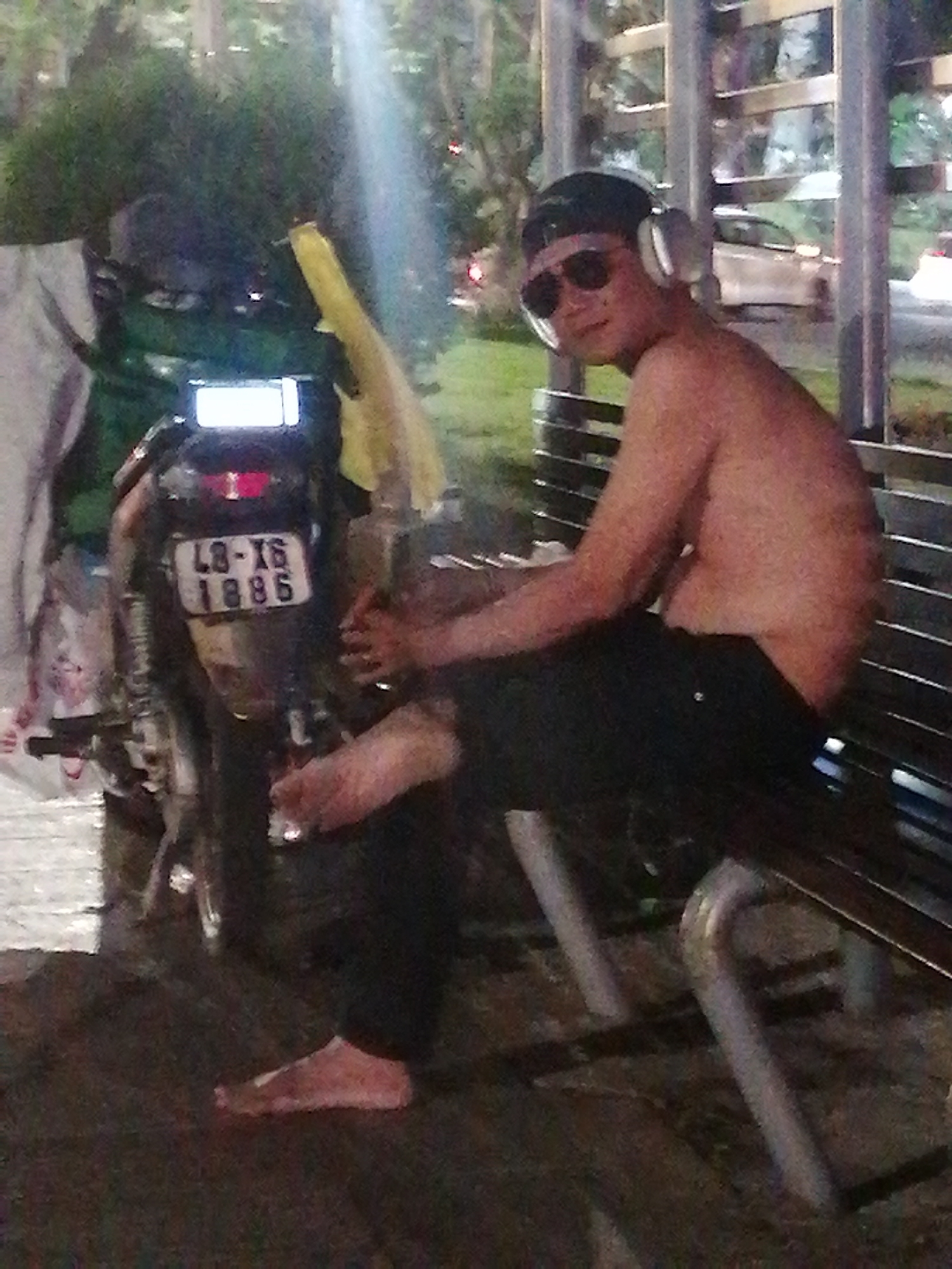 A shirtless man sits on a bench in Da Lat, Vietnam, next to a motorcycle. The man is wearing headphones, sunglasses, and a baseball cap. He is barefoot and has black pants on. The license plate of the motorcycle reads "49-X6 1886." The background includes trees, street lights, and a car.