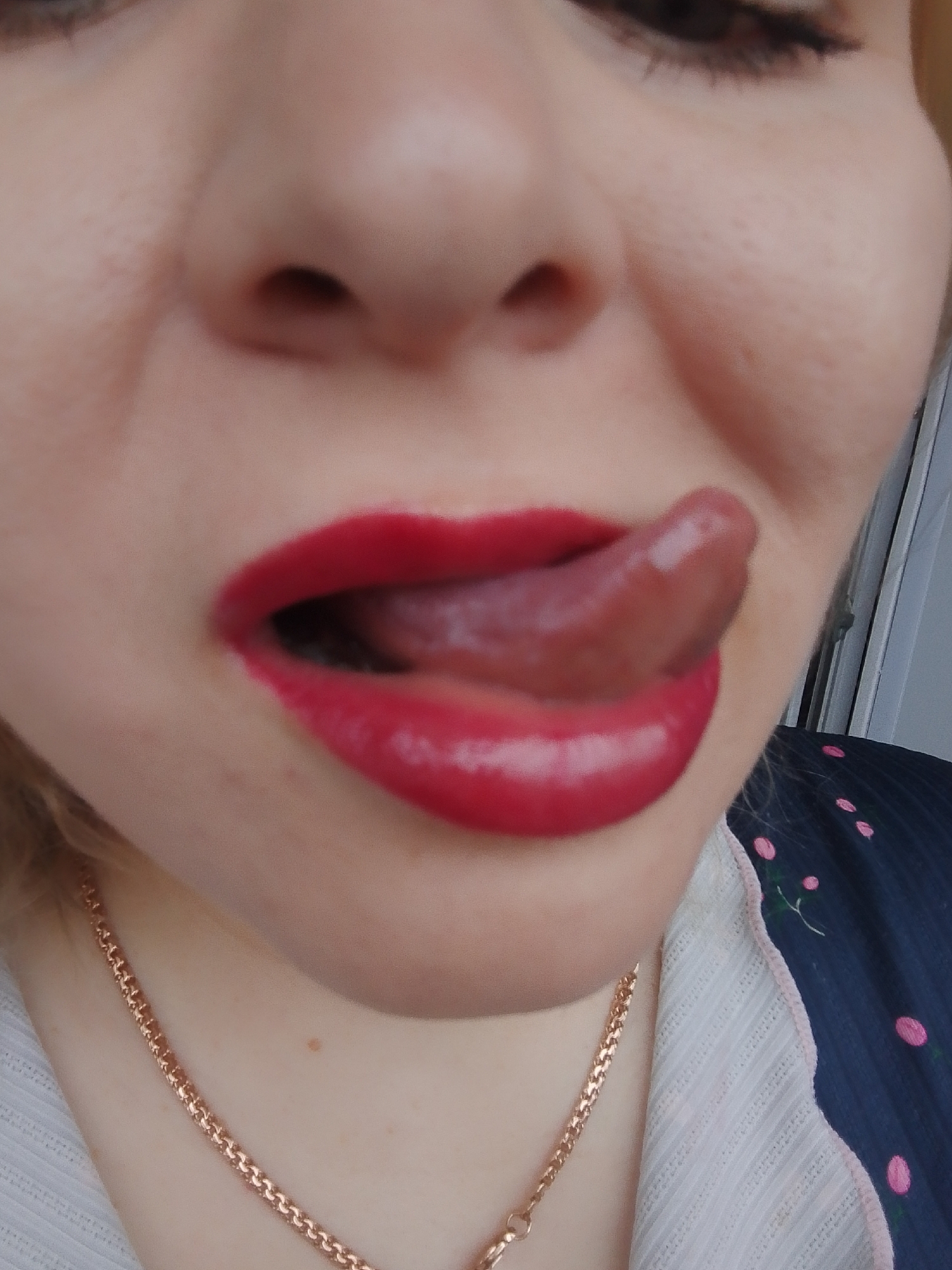 A close-up, high-angle photograph depicts a portion of a person's face. The subject is fair-skinned. The person's mouth is open, with the tongue protruding outward and slightly upward. The lips are coated in bright red lipstick. Parts of the nose and one eye are visible, indicating the focus is on the mouth. Below the chin, there is a gold chain necklace. The person is wearing a shirt that has a pink floral design on a dark background. The location is Izhevsk, Russia.