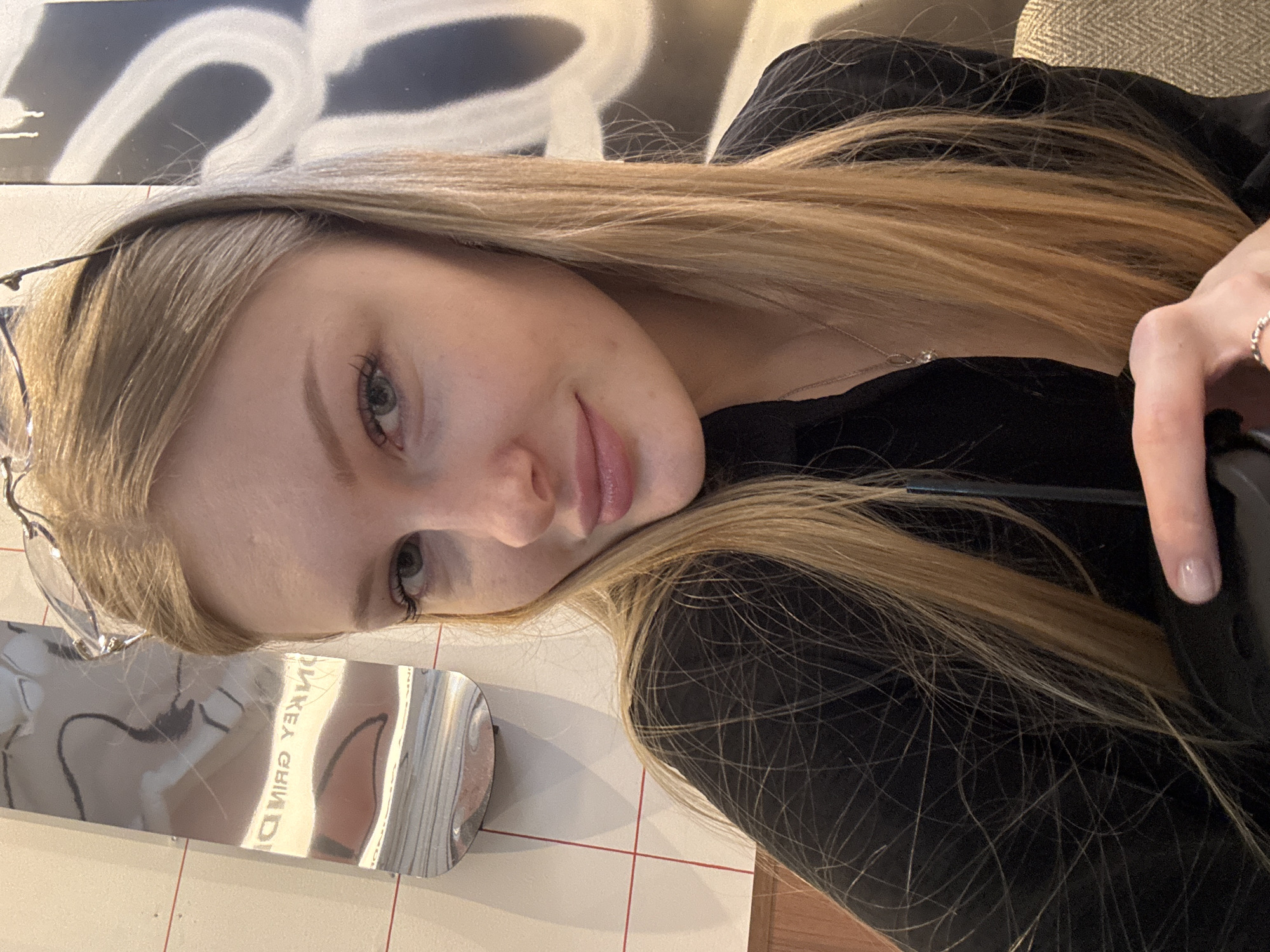 A fair-skinned woman with long blonde hair, wearing a black top and a silver necklace, is facing the camera. She is holding a dark-colored object, possibly a phone, with her right hand. The image is taken indoors, with a reflective silver object on a tiled surface to the left. There is some writing visible in the reflection of the silver object, reading "...MKEA GRIND". The background features a white and grey graffiti pattern. The location is in Perm, Russia.