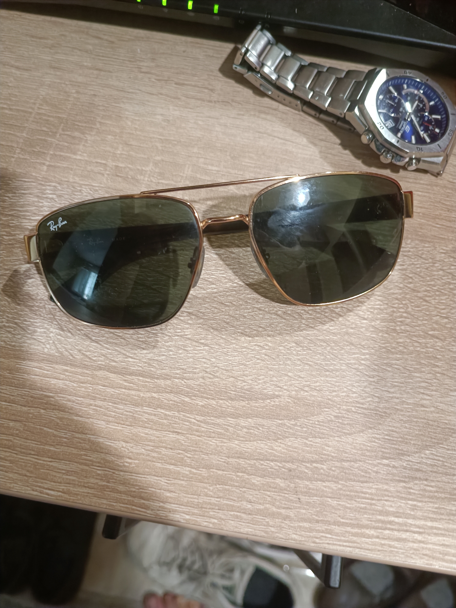 In Burgas, Bulgaria, a pair of gold-rimmed Ray-Ban sunglasses with dark lenses lies on a light-colored wooden surface. To the upper right of the sunglasses, a silver-banded watch with a blue face is visible, also placed on the same wooden surface. A dark, flat object, possibly a keyboard, is partially visible at the top of the image. The lighting suggests an indoor setting.