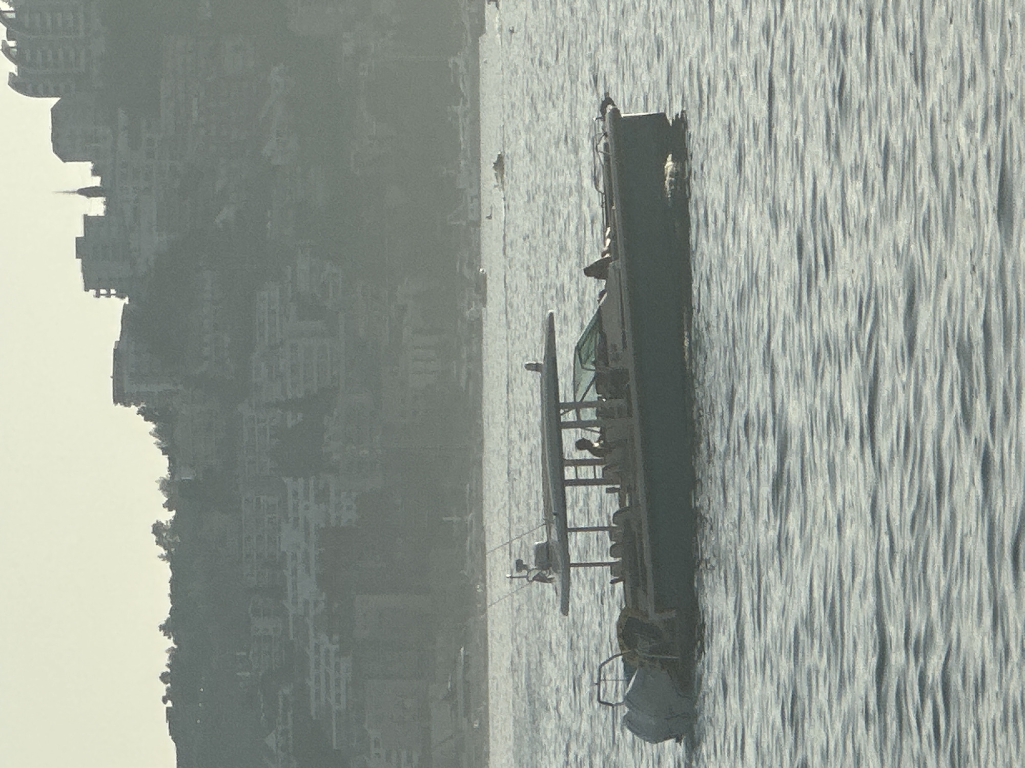 A boat with a canopy is on the water, with a city built into a hillside in the background. The boat has a light-colored hull and a dark-colored canopy supported by poles. There are two people visible on the boat, one appearing to be at the helm. The water is choppy with moderate waves. The background shows a densely built hillside with numerous buildings, suggesting a coastal city. The overall lighting is hazy, indicative of either early morning, late afternoon, or atmospheric conditions like fog or smog. The architecture in the background, particularly the layered development on the hillside, is characteristic of the coastal areas of Budva, Montenegro, which is known for its hilly terrain and dense urban development along the Adriatic Sea.