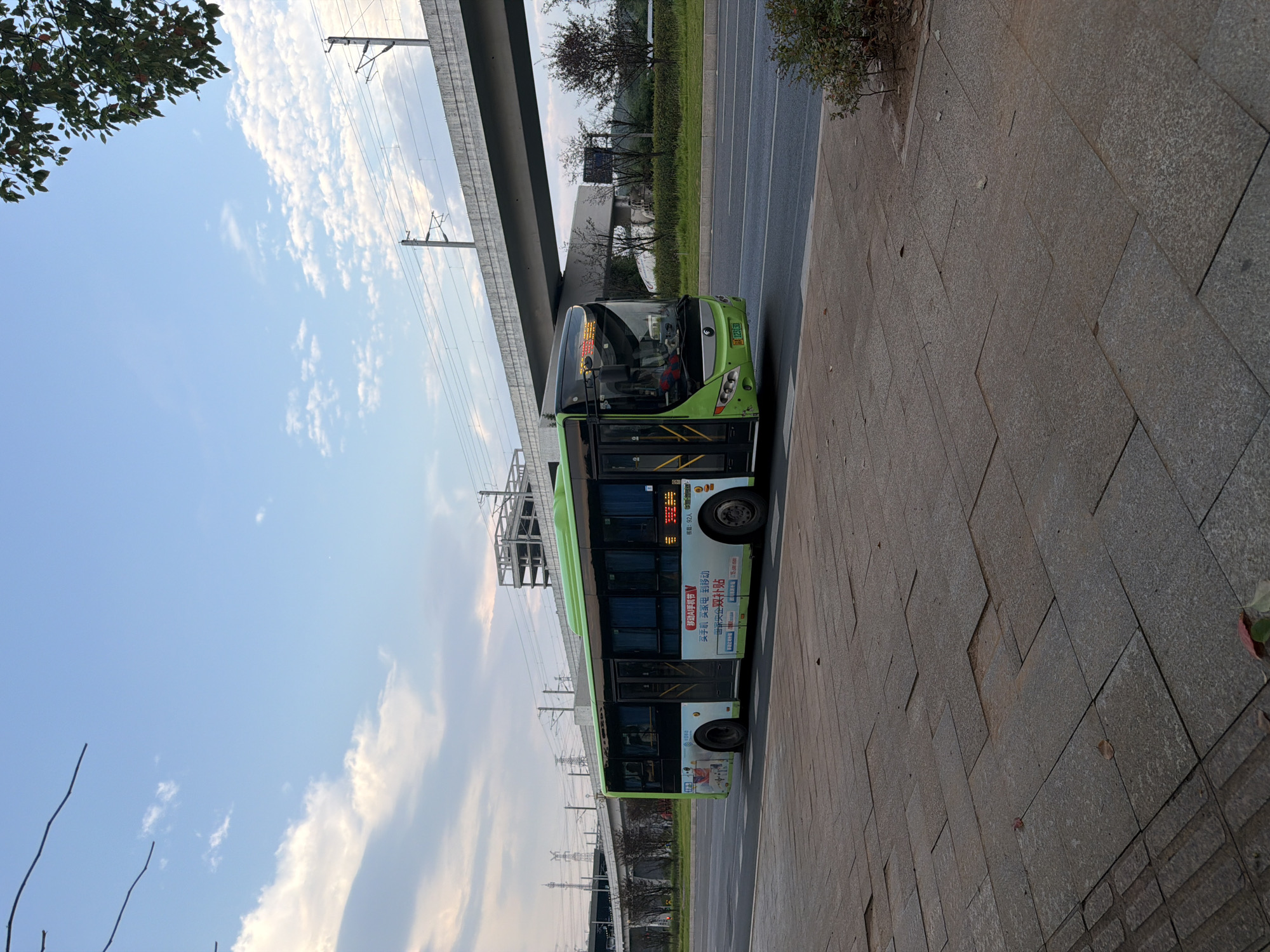 A green and white bus is driving down a city street in Jiangjin, China. The bus has advertisements on its side and a route number displayed on its front. In the background, there is an elevated railway line and some buildings. The sky is blue with some clouds. The street is paved with concrete tiles.