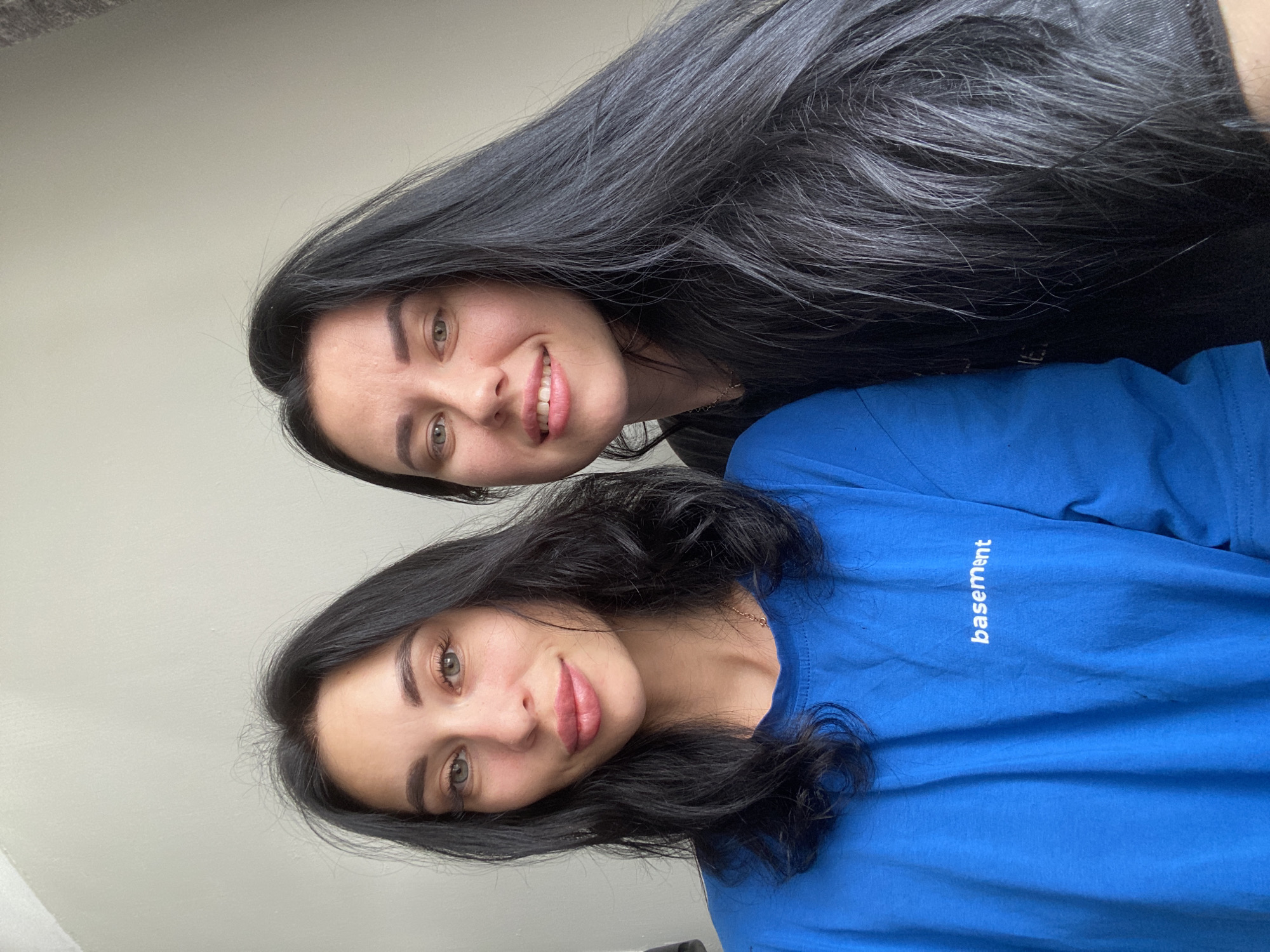 The image is a selfie taken indoors, featuring two women with dark hair against a plain, light-colored wall. The woman on the left, closer to the camera, has medium-length wavy hair and is wearing a bright blue t-shirt with the word "basement" printed in white on the chest. She is looking directly at the camera with her mouth slightly open, revealing her teeth. The woman on the right has long, straight dark hair and is also looking towards the camera, smiling. She is wearing a dark t-shirt. The lighting appears to be natural, coming from the front. The image does not provide any specific details that would allow for the determination of its location within the city of Chaikovsky, Russia.