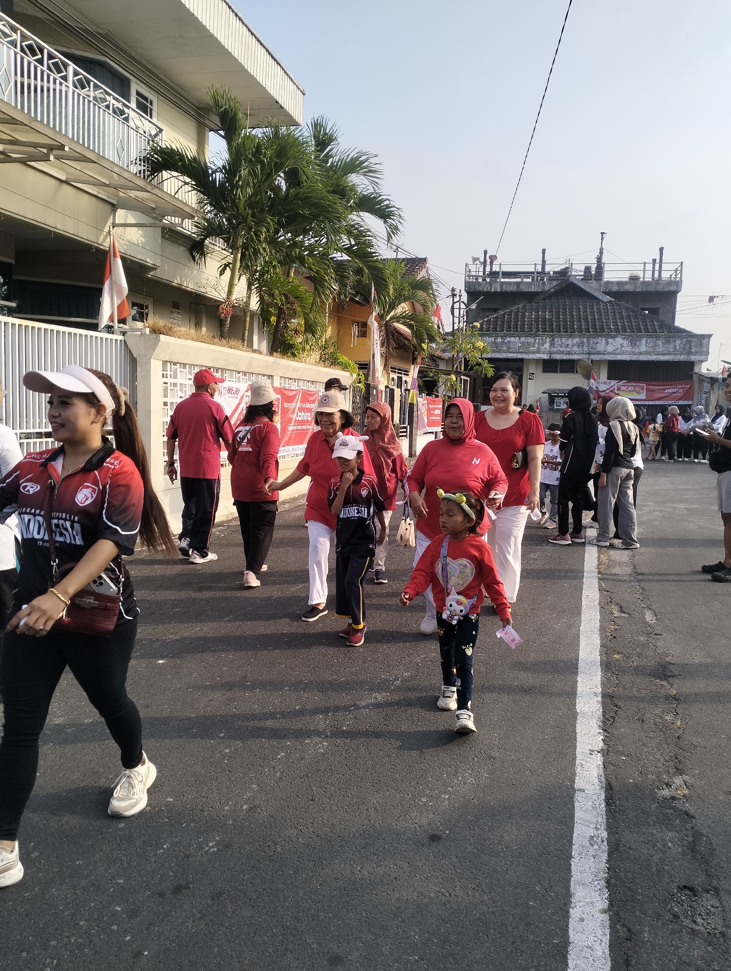 The image captures a street scene in the city of Magelang, Indonesia, likely during an outdoor event or procession. A group of people, predominantly women and children, are walking on a paved road. Many are wearing red shirts, some with the text "INDONESIA" visible. The architecture in the background suggests a mix of residential and commercial buildings, with a prominent building on the left featuring a white fence and balconies. Palm trees are interspersed, indicating a tropical climate. Red and white Indonesian flags are displayed on some of the buildings. The atmosphere appears to be lively and communal, with people engaged in walking and interacting. The lighting suggests it is daytime, likely morning or late afternoon due to the angle of the sun. The road surface shows signs of wear and tear, with a white line marking the lane.