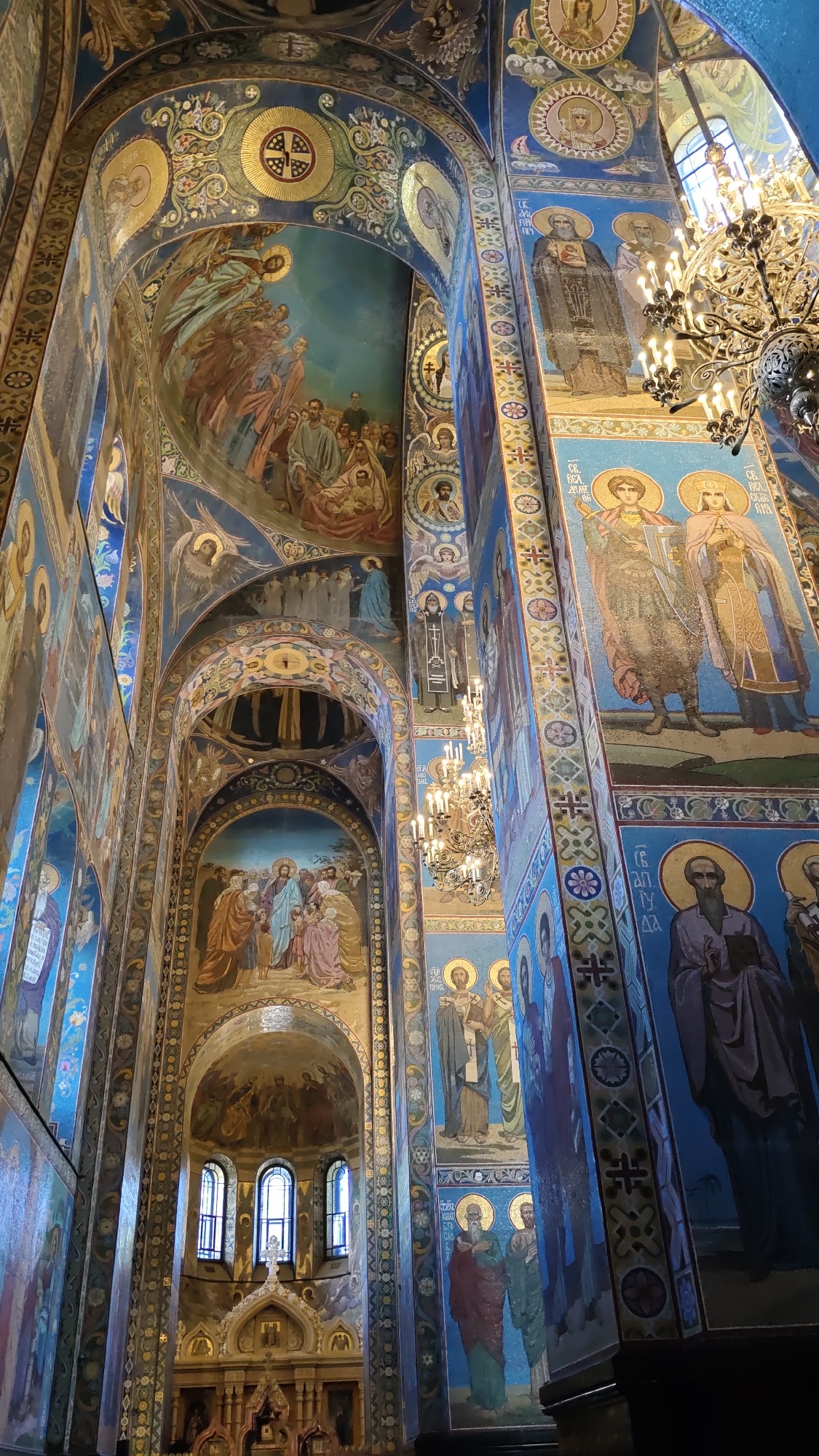 This image shows the interior of a richly decorated cathedral, likely in Saint Petersburg, Russia. The walls and ceilings are adorned with numerous religious mosaics and paintings depicting various biblical scenes and figures, including what appears to be the Nativity on the left arch and Jesus with followers on the lower central arch. Stylized figures of saints and religious symbols are meticulously rendered in vibrant blues, golds, and other colors.
A large, ornate chandelier with multiple candle-like lights hangs from the ceiling, casting a warm glow and illuminating the intricate details of the mosaics. The architecture features grand arches and columns, typical of a large religious structure. The lighting suggests it is daytime, with light filtering in from windows that are visible in the background, though the interior itself is dimly lit by the chandelier. The overall impression is one of solemnity and grandeur, highlighting the religious and artistic significance of the location. No visible text is discernible in the image.