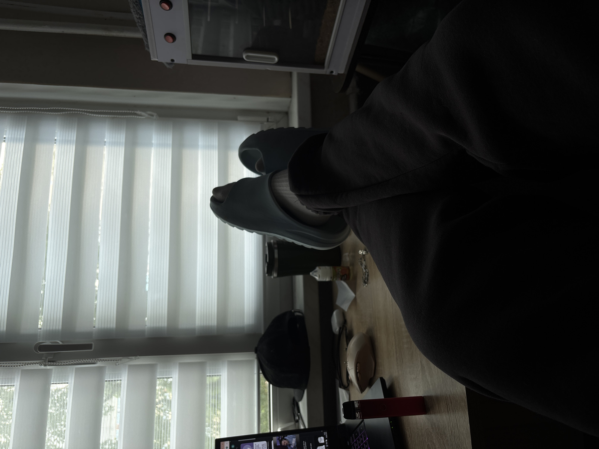 The image depicts a person's legs and feet, clad in dark pants and light blue slides, resting on a desk in a dimly lit room. The desk is made of wood and has various objects on it, including a laptop, a red vape pen, a light-colored computer mouse, a black baseball cap, a dark thermos, a small bottle of liquid, and a silver chain. Behind the desk, a window is covered by horizontal blinds, allowing diffused light to enter the room. To the right of the window, a white appliance with red lights and a glass door is visible. The overall scene suggests a casual or work-from-home environment. There is no information in the image to indicate the location in the city of Saint Petersburg, Russia.
