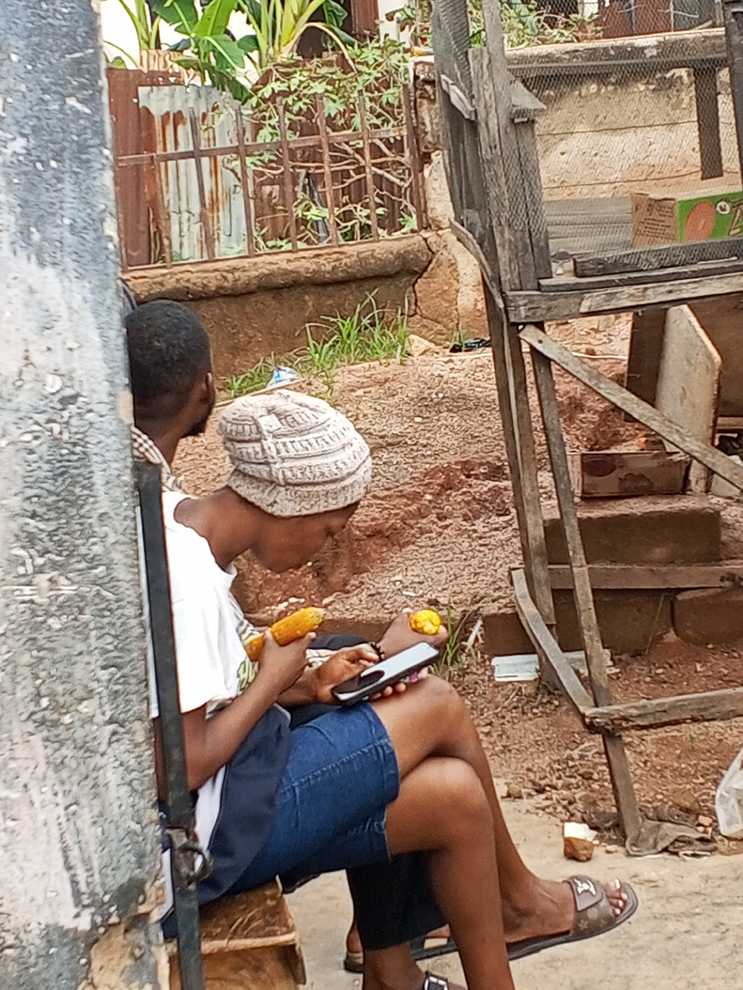 A photo taken in Gwong, Nigeria, shows two individuals seated in an outdoor urban setting. The person in the foreground is wearing a white t-shirt, blue shorts, and a light-colored knitted hat. This person is holding a piece of corn in one hand and a smartphone in the other, looking down at the phone. A second individual sits beside the first, mostly obscured except for their head and the upper portion of their back. A concrete wall with peeling paint is visible to the left of the frame. Behind the subjects, there is a raised dirt area with vegetation, along with a weathered wooden structure that resembles stairs or scaffolding. Debris is scattered on the ground. A metal fence and additional foliage can be seen further in the background.